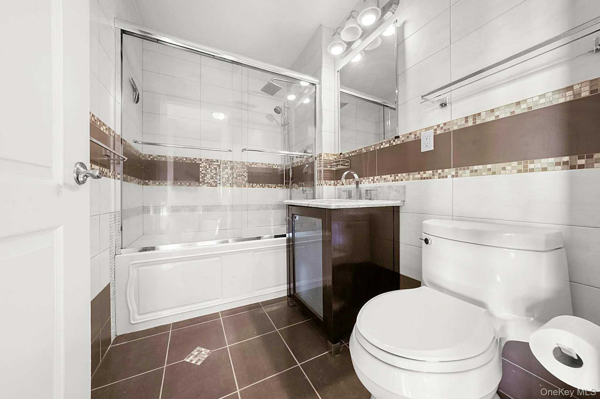 Bathroom featuring a white bathtub with a clear glass sliding shower door, a dark wood-finish vanity with a white countertop and undermount sink, a large mirror with a four-bulb vanity light, and brown floor tiling with decorative mosaic accents Bathroom featuring a white bathtub with a clear glass sliding shower door, a dark wood-finish vanity with a white countertop and undermount sink, a large mirror with a four-bulb vanity light, and brown floor tiling with decorative mosaic accents