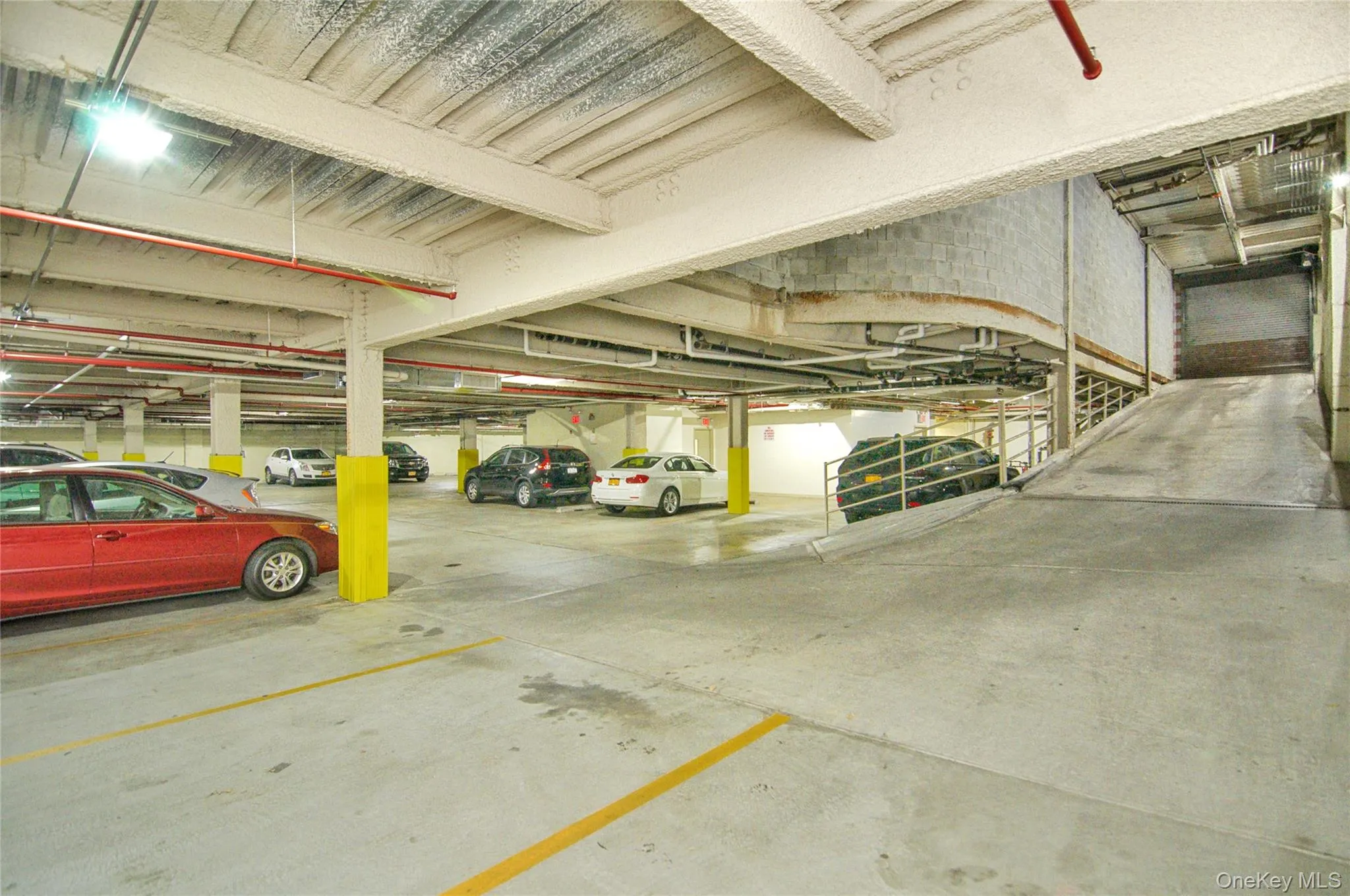 Underground parking garage featuring concrete flooring, structural support columns, overhead lighting, and an access ramp Underground parking garage featuring concrete flooring, structural support columns, overhead lighting, and an access ramp