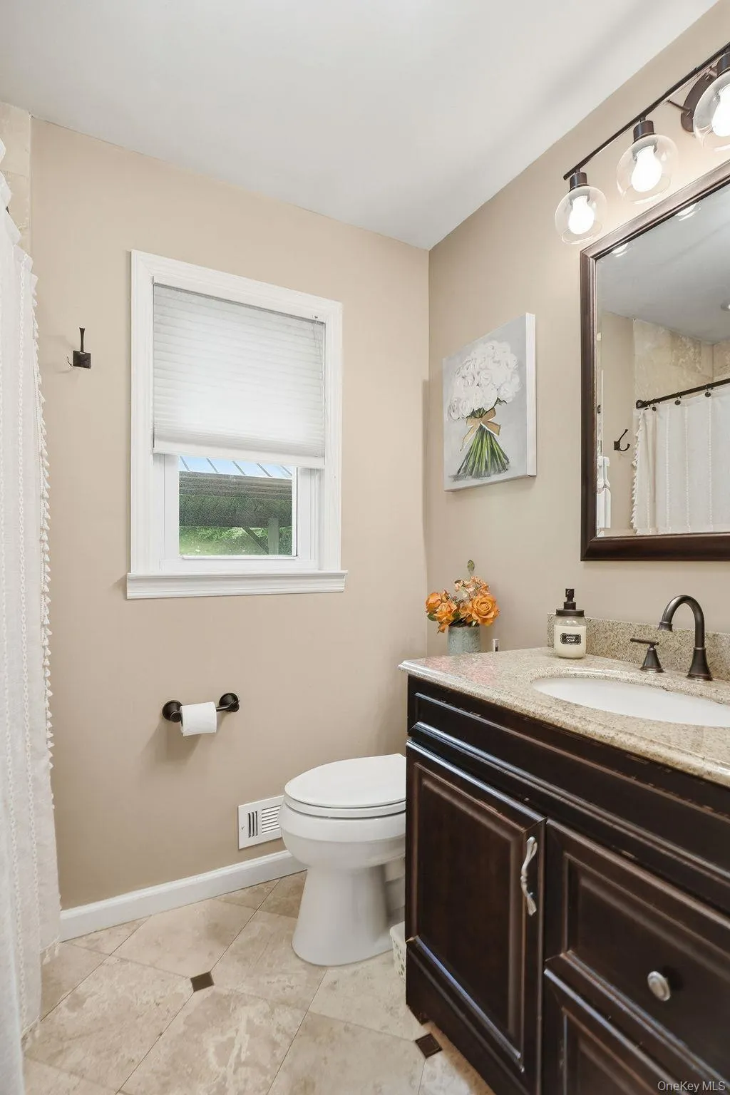 Bathroom featuring a dark wood vanity with a light-toned countertop, a rectangular mirror with a three-light fixture, and a window with white trim Bathroom featuring a dark wood vanity with a light-toned countertop, a rectangular mirror with a three-light fixture, and a window with white trim