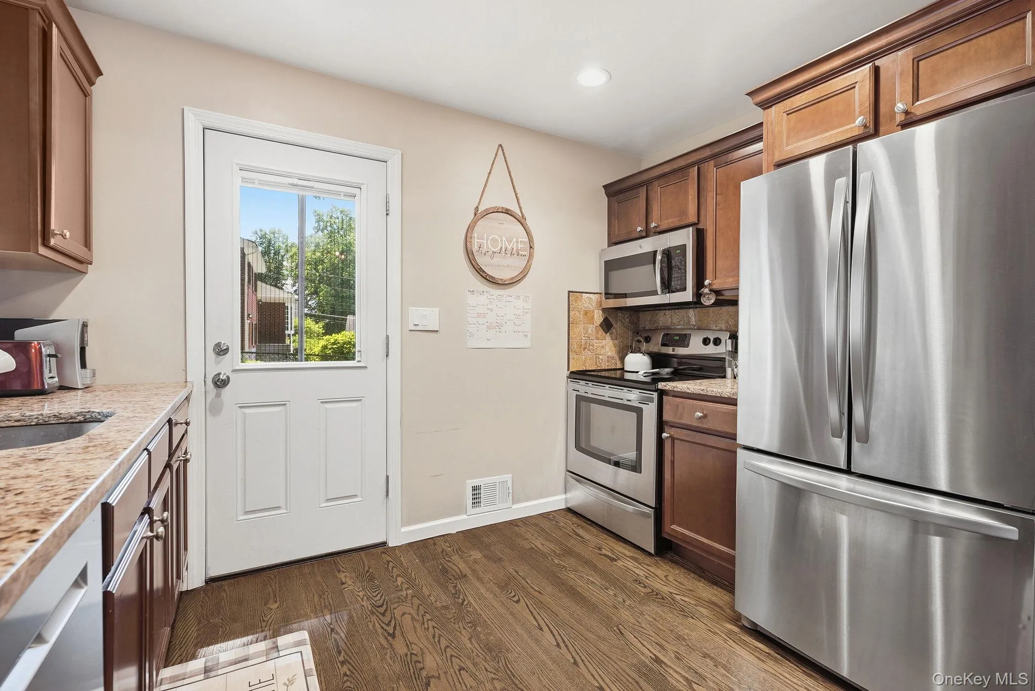 Well-appointed kitchen featuring wood-finish flooring, granite-style countertops, stainless steel appliances, dark wood cabinetry, and a glass-paneled door Well-appointed kitchen featuring wood-finish flooring, granite-style countertops, stainless steel appliances, dark wood cabinetry, and a glass-paneled door