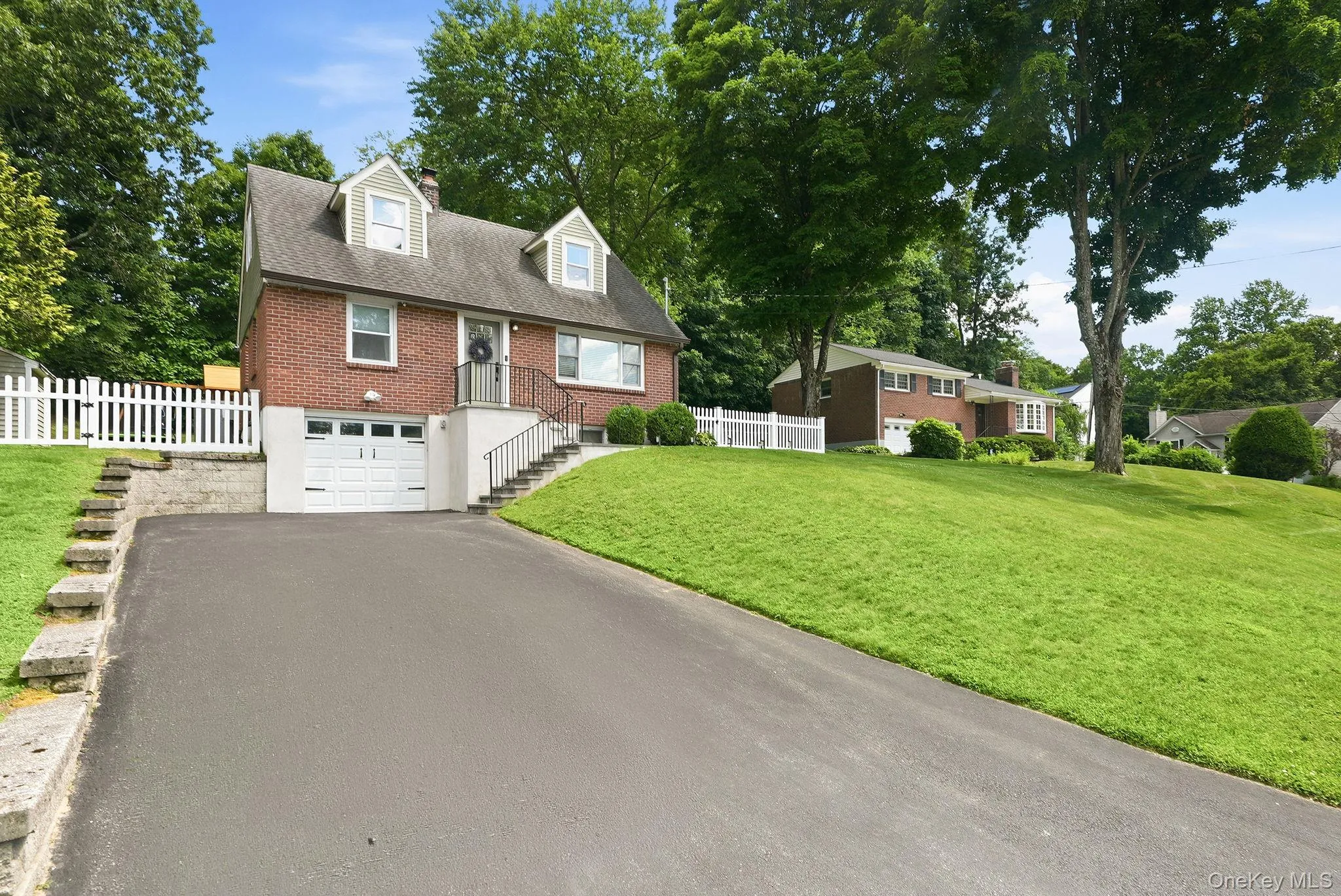 Brick-front residence featuring a dormered roofline, white picket fence, and an attached garage with a paved driveway Brick-front residence featuring a dormered roofline, white picket fence, and an attached garage with a paved driveway