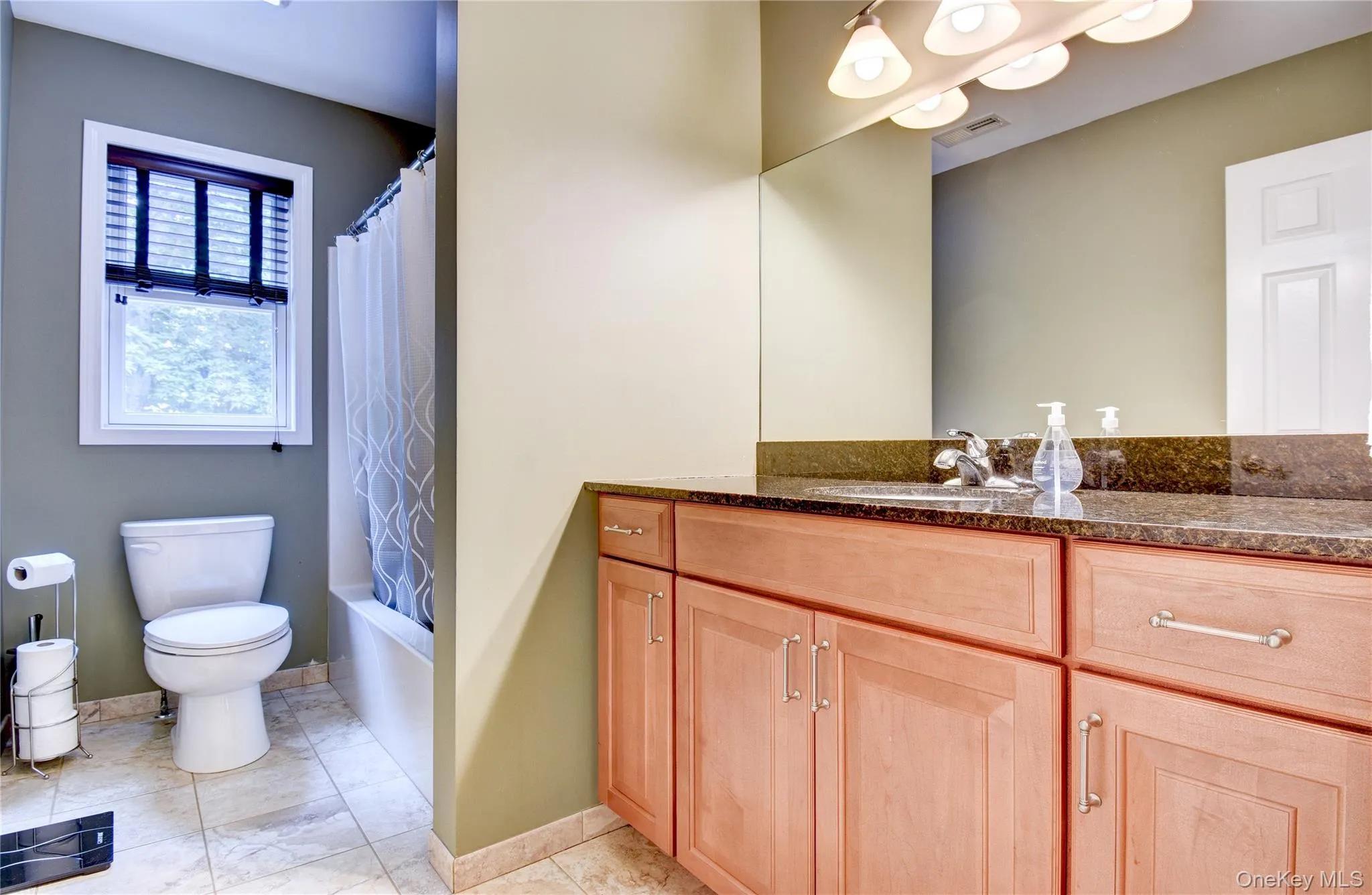 Hall Bathroom featuring a wood-finish vanity with a dark granite countertop and integrated sink, a large wall-mounted mirror, and a sconce light fixture Hall Bathroom featuring a wood-finish vanity with a dark granite countertop and integrated sink, a large wall-mounted mirror, and a sconce light fixture