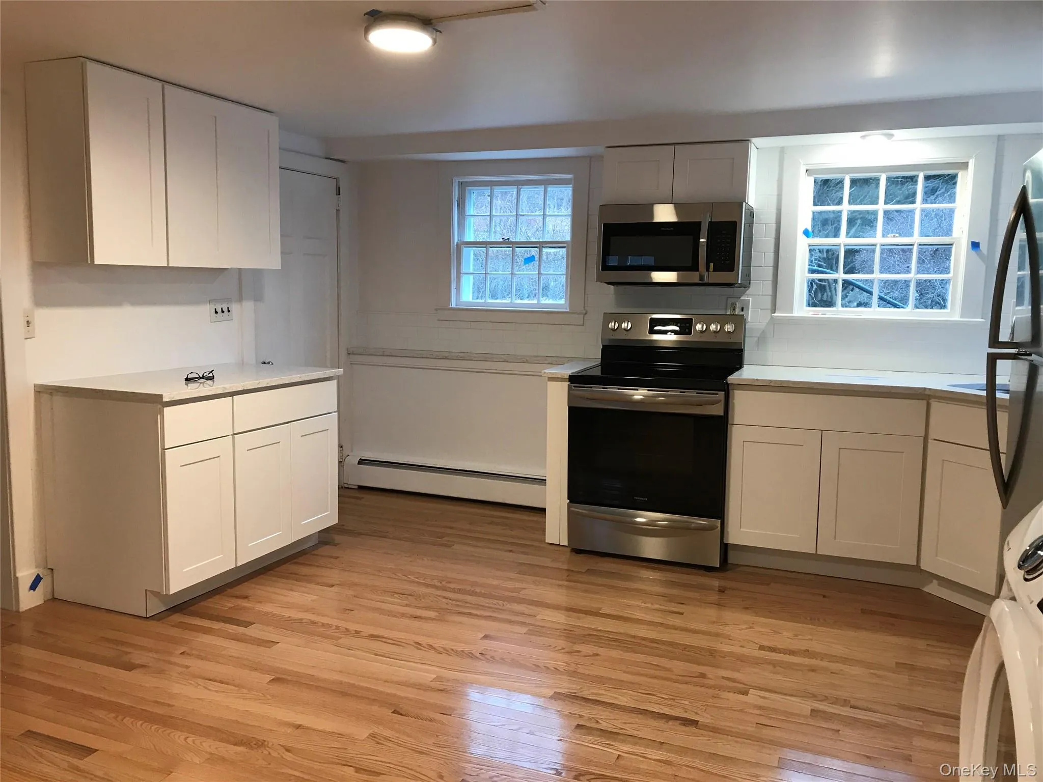 Bright kitchen featuring hardwood flooring, white shaker cabinetry, light-toned countertops, and stainless steel appliances Bright kitchen featuring hardwood flooring, white shaker cabinetry, light-toned countertops, and stainless steel appliances
