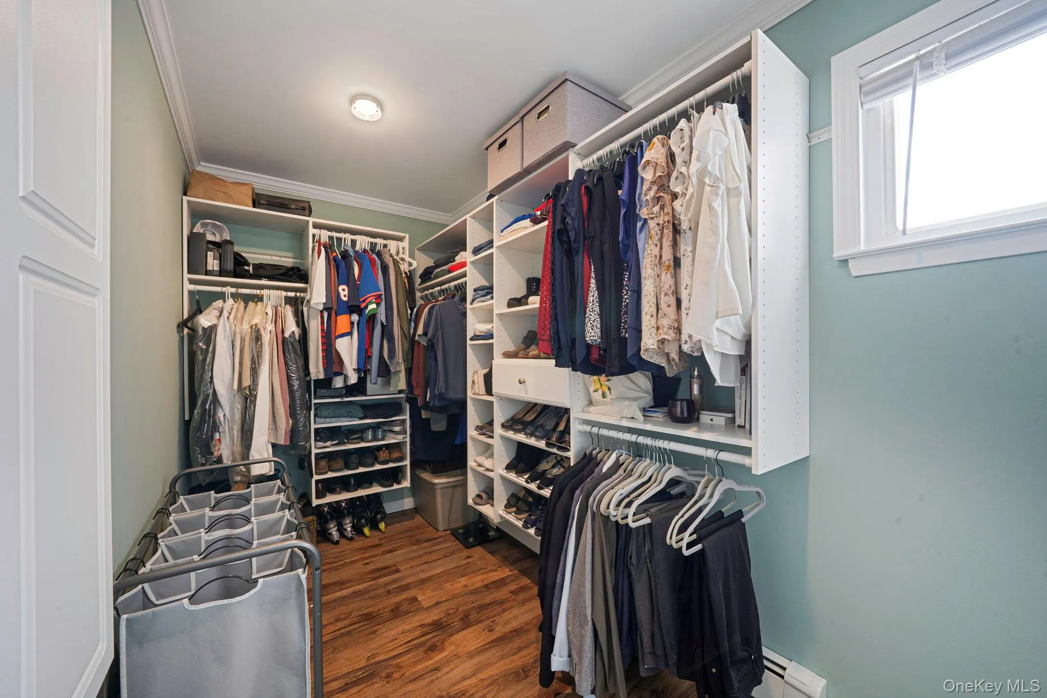 Walk-in closet featuring custom built-in shelving, multiple hanging rods, dedicated shoe storage, wood-finish flooring, and natural light from a interior window Walk-in closet featuring custom built-in shelving, multiple hanging rods, dedicated shoe storage, wood-finish flooring, and natural light from a interior window