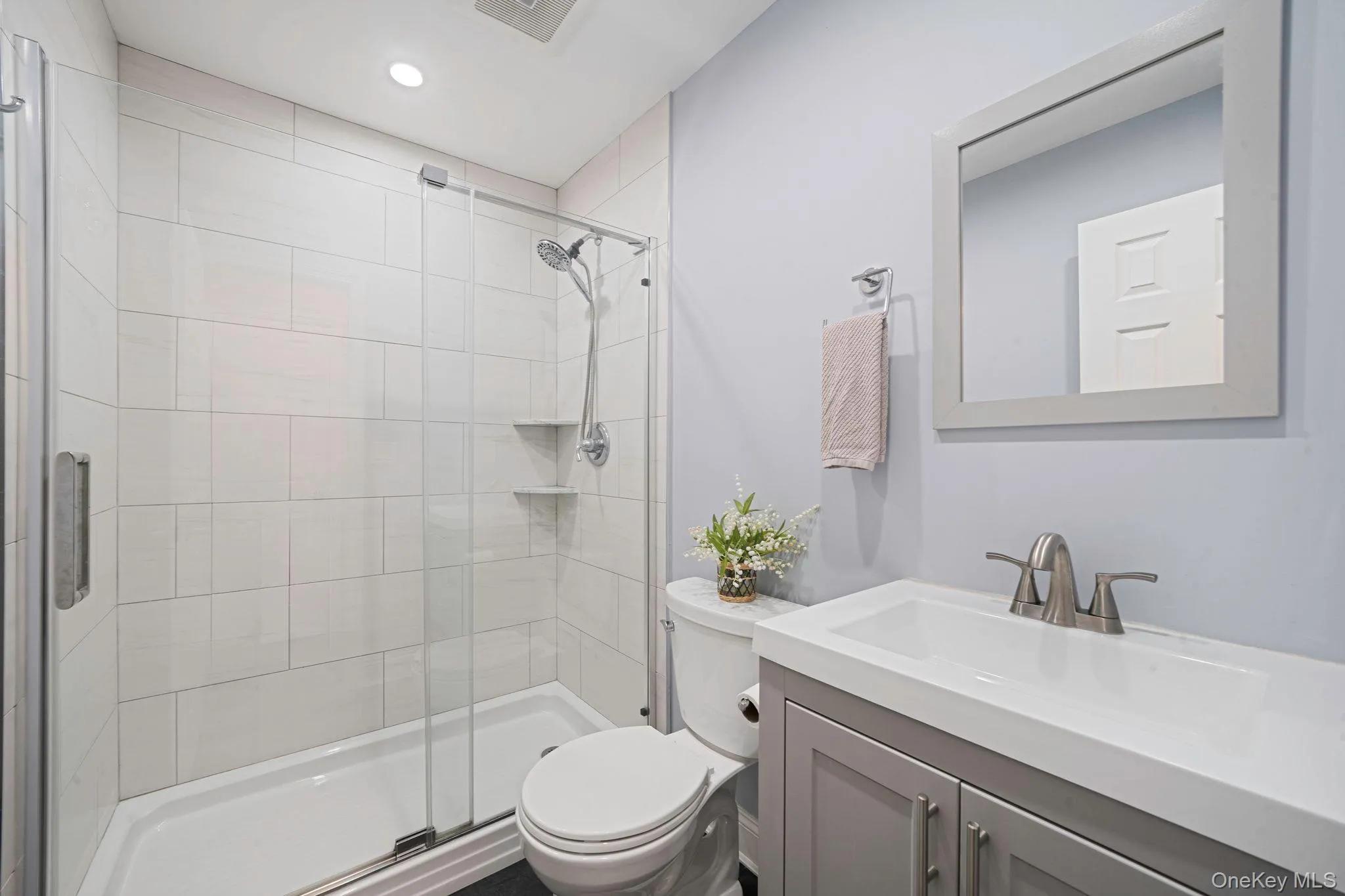1st floor full Bathroom featuring a glass-enclosed shower with white tiled walls and built-in shelving, a gray vanity with an integrated white sink, a brushed nickel faucet, and a classic framed mirror 1st floor full Bathroom featuring a glass-enclosed shower with white tiled walls and built-in shelving, a gray vanity with an integrated white sink, a brushed nickel faucet, and a classic framed mirror