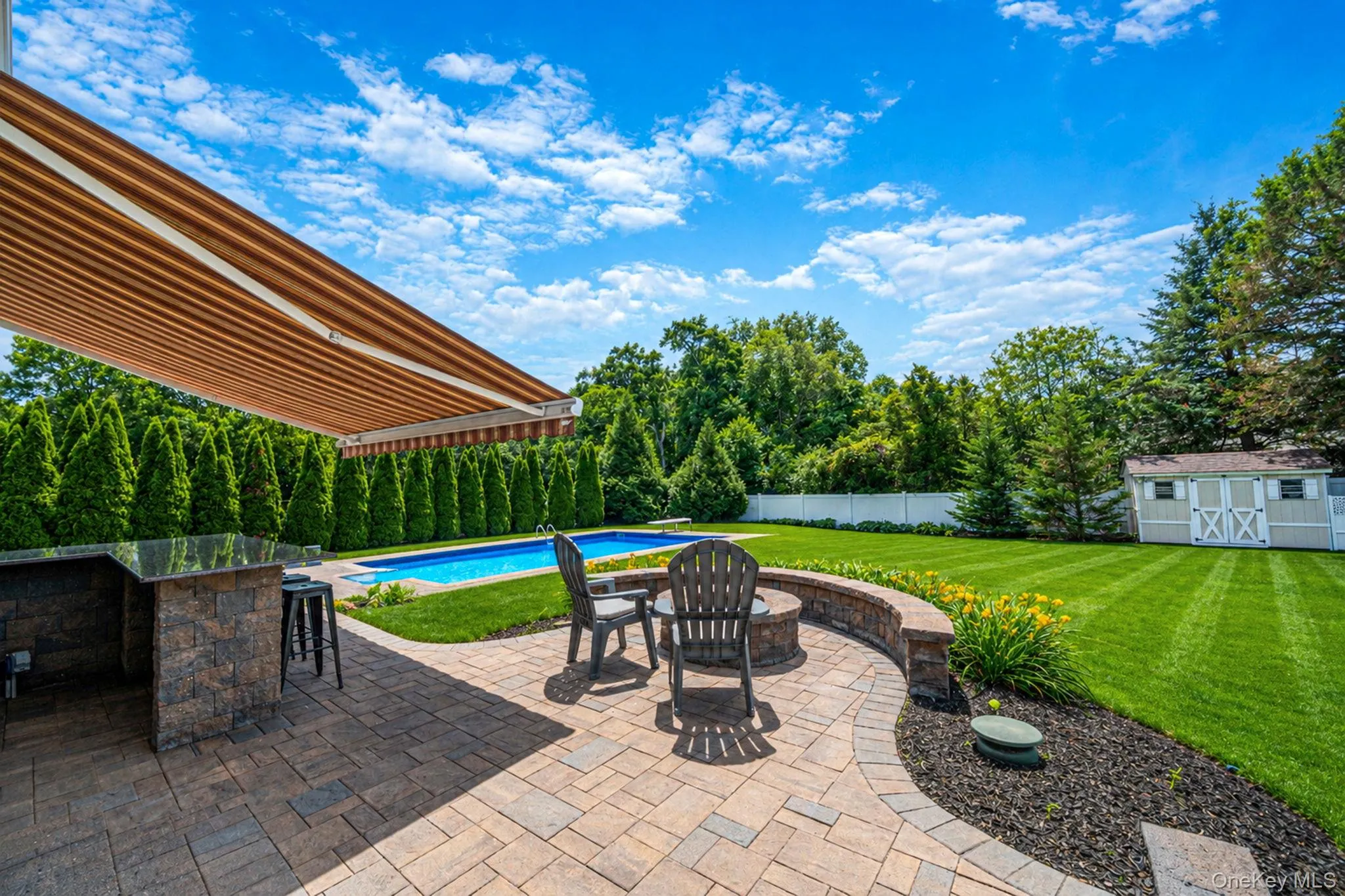 Spacious outdoor living area with a built-in stone granite island, retractable awning, paver patio, in-ground pool, and a fire pit. In ground sprinkler system. Large Shed for additional storage. Spacious outdoor living area with a built-in stone granite island, retractable awning, paver patio, in-ground pool, and a fire pit. In ground sprinkler system. Large Shed for additional storage.