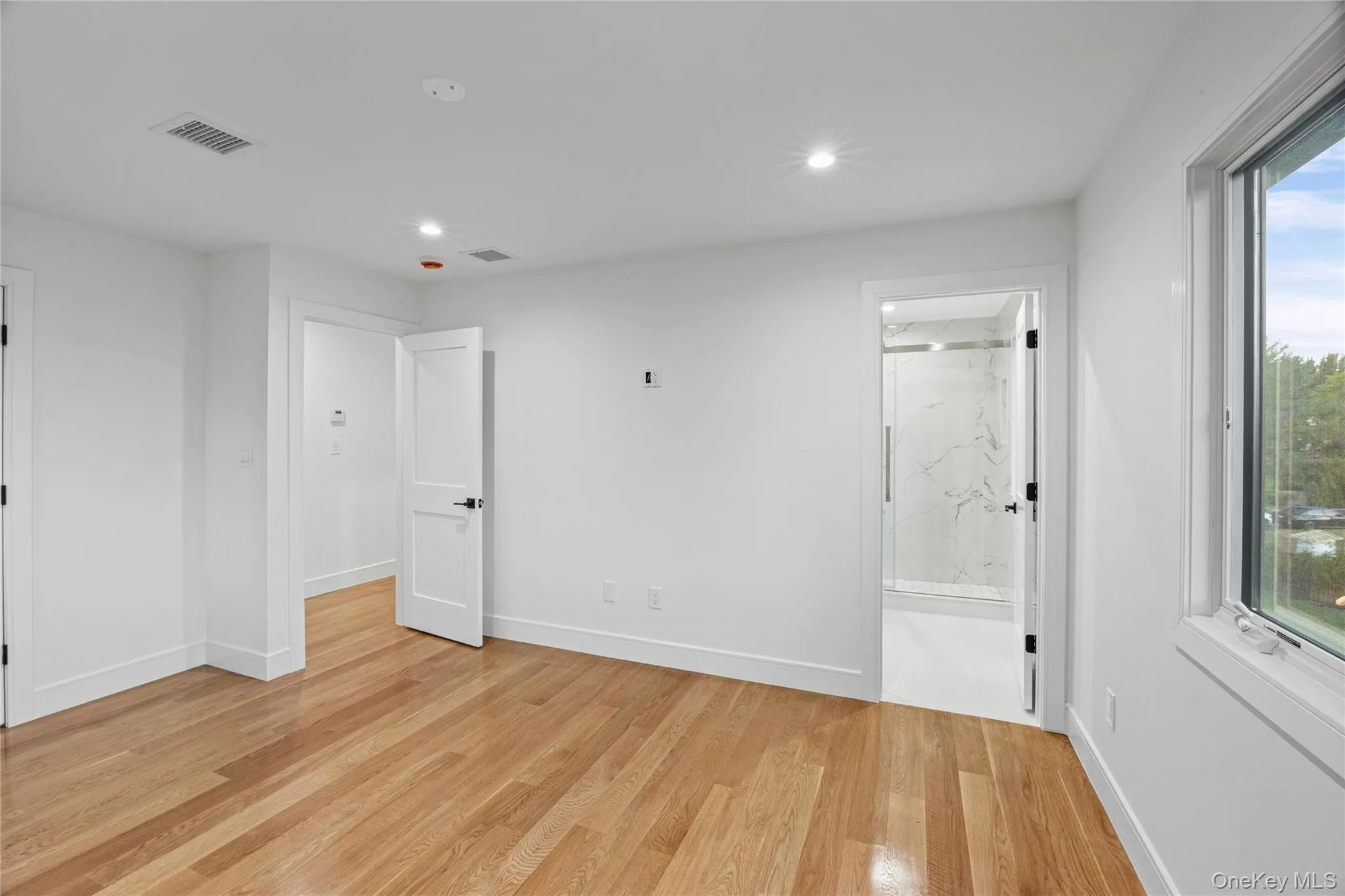 Spacious bedroom with en-suite bathroom, featuring wood flooring, neutral wall tones, recessed lighting, and a window providing natural light. Spacious bedroom with en-suite bathroom, featuring wood flooring, neutral wall tones, recessed lighting, and a window providing natural light.