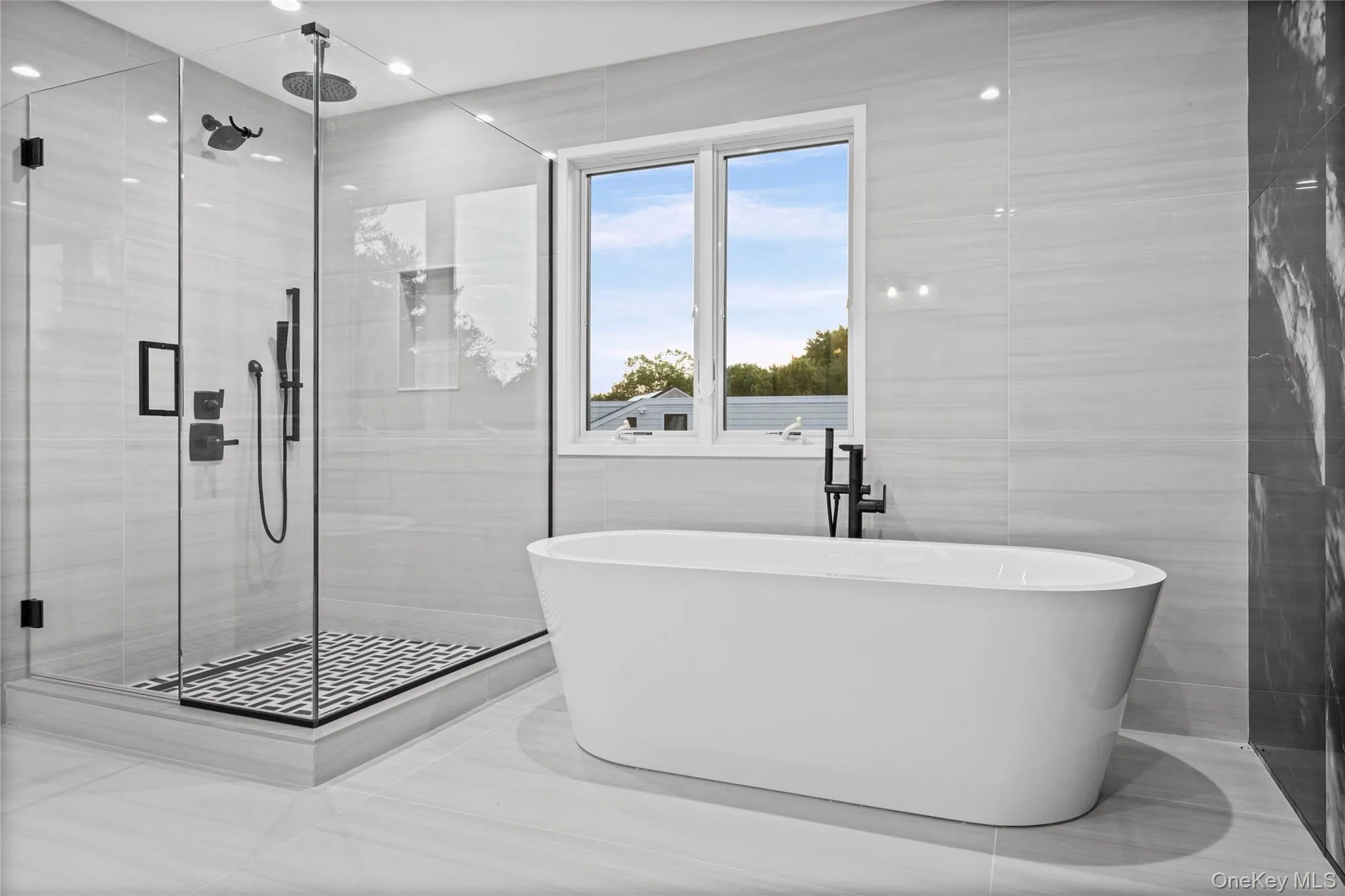 Bathroom featuring a freestanding soaking tub with floor-mounted faucet, a glass-enclosed shower with a rainfall showerhead and hand shower, and light-toned tile flooring and walls Bathroom featuring a freestanding soaking tub with floor-mounted faucet, a glass-enclosed shower with a rainfall showerhead and hand shower, and light-toned tile flooring and walls