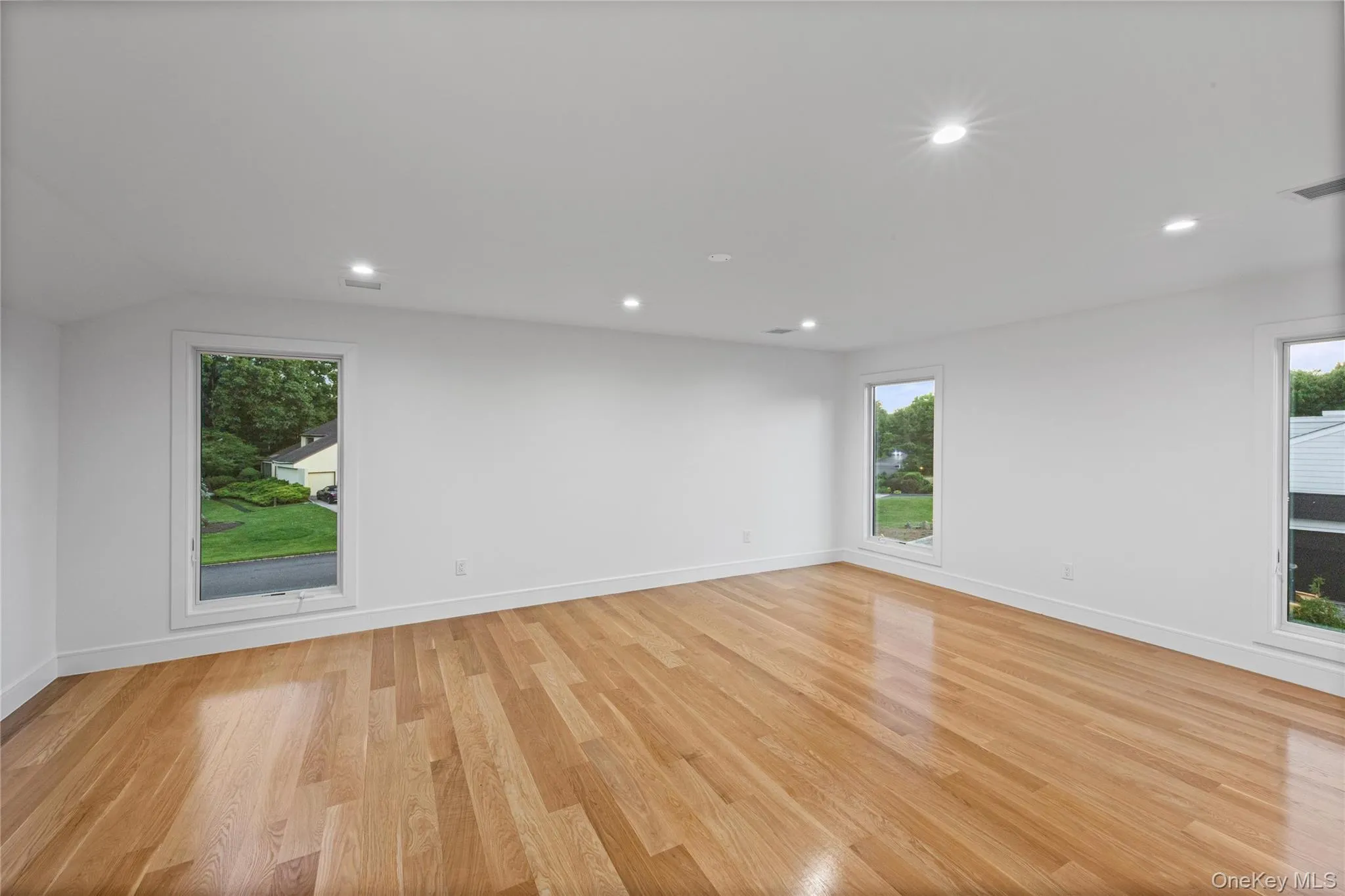 Spacious room featuring hardwood flooring, multiple windows with white trim, recessed lighting, and a neutral color palette Spacious room featuring hardwood flooring, multiple windows with white trim, recessed lighting, and a neutral color palette