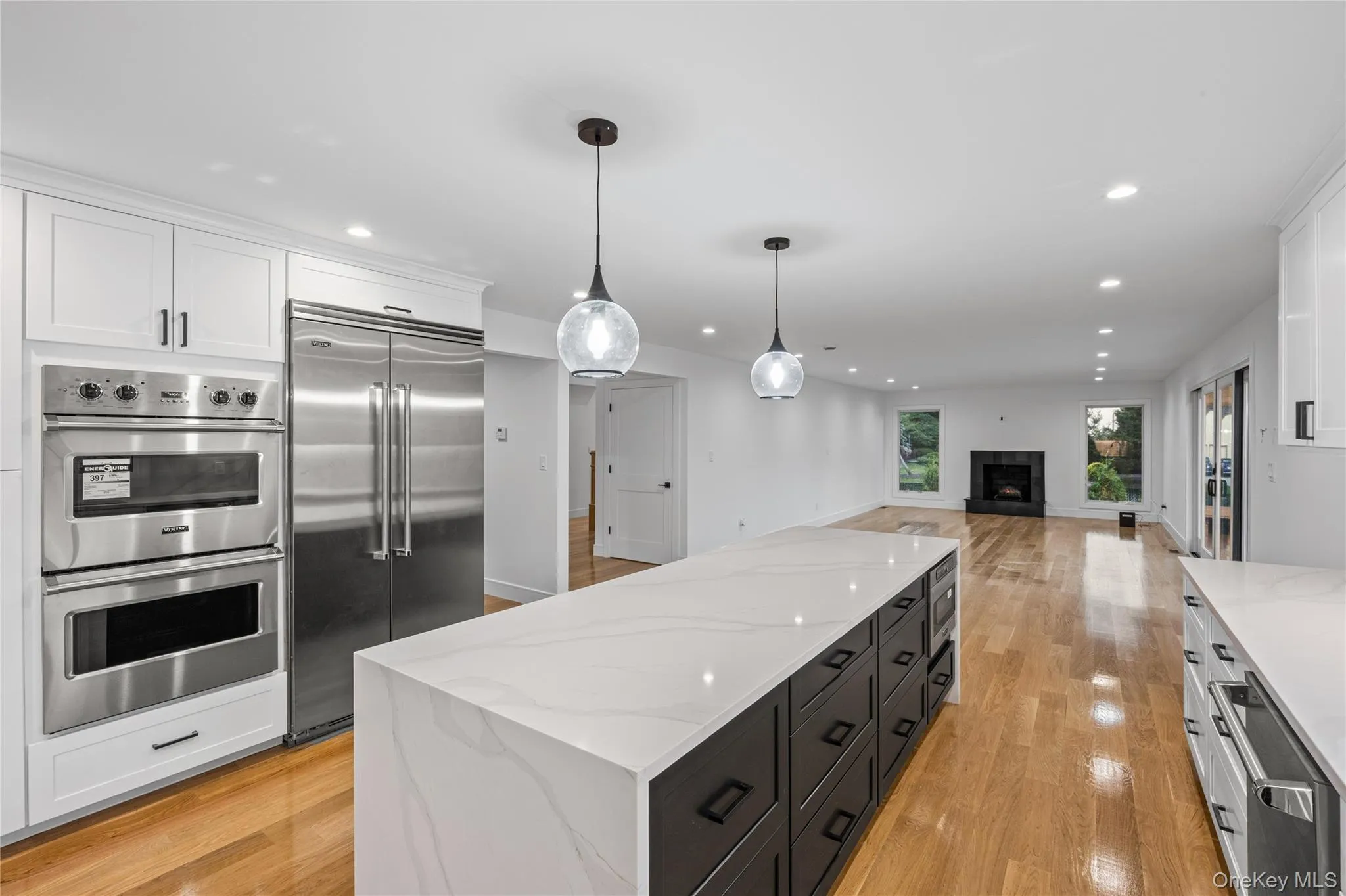 Modern kitchen featuring an expansive island with a waterfall countertop, white cabinetry, integrated stainless steel appliances, and wood-finish flooring. Viking Appliances. Modern kitchen featuring an expansive island with a waterfall countertop, white cabinetry, integrated stainless steel appliances, and wood-finish flooring. Viking Appliances.