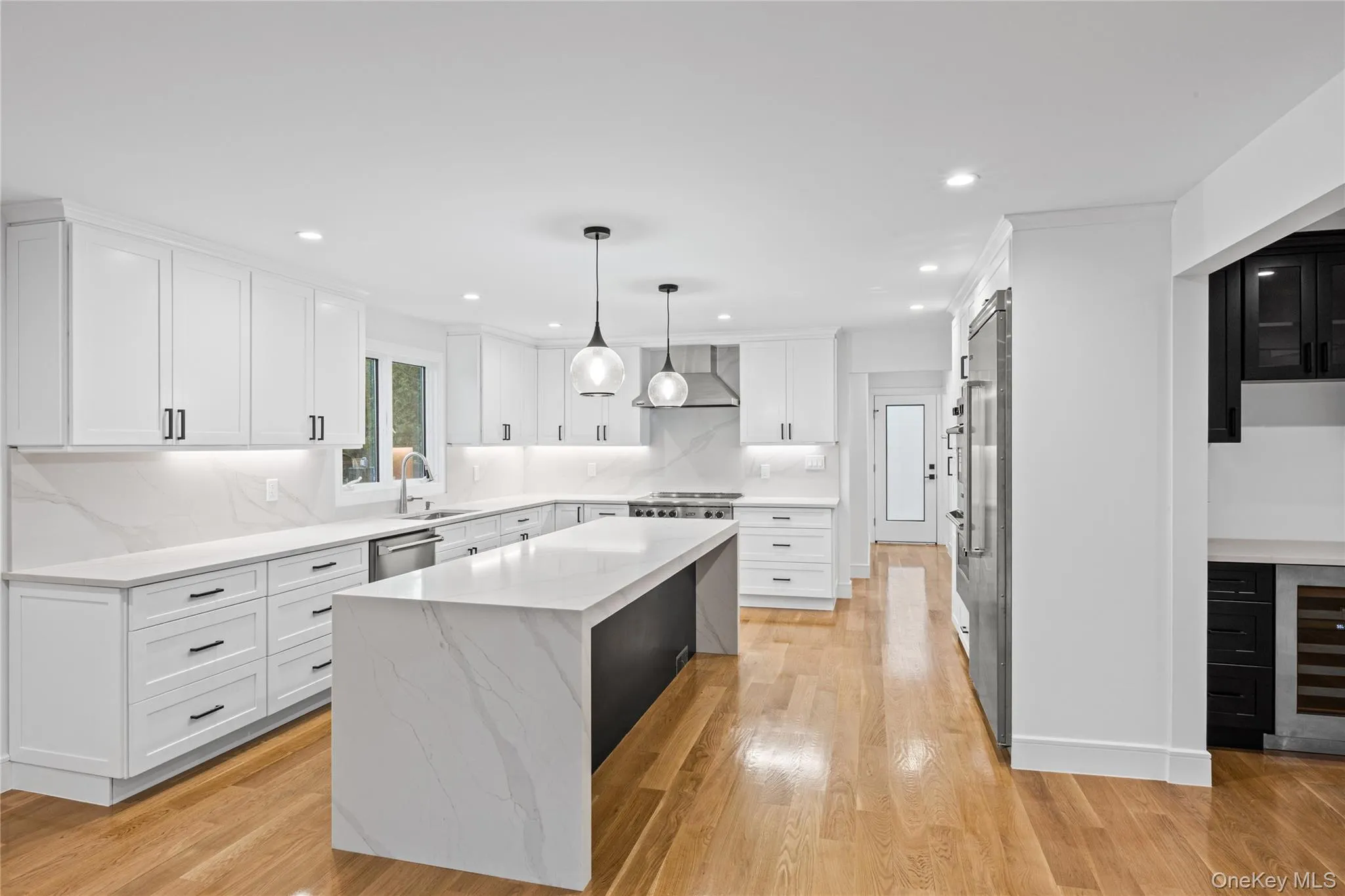 Bright kitchen featuring white cabinetry, a large center island with waterfall countertop, stainless steel appliances, and wood-finish flooring Bright kitchen featuring white cabinetry, a large center island with waterfall countertop, stainless steel appliances, and wood-finish flooring