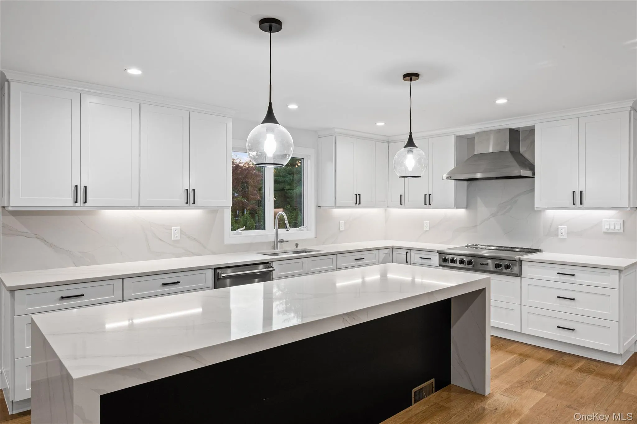 Modern kitchen featuring a waterfall island with a quartz countertop, white shaker-style cabinetry with matte black hardware, and wood-finish flooring Modern kitchen featuring a waterfall island with a quartz countertop, white shaker-style cabinetry with matte black hardware, and wood-finish flooring