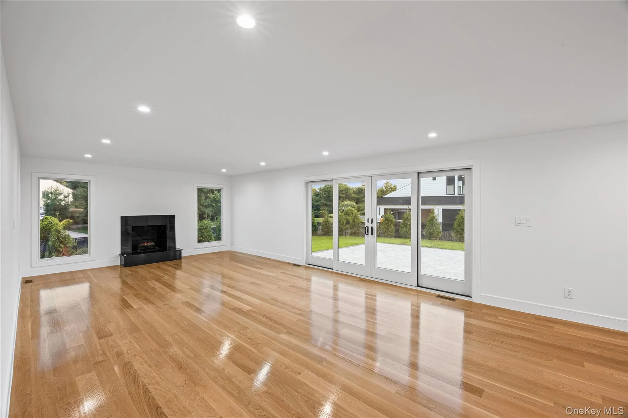 Spacious room featuring wood-finish flooring, a black stone fireplace, recessed lighting, two rectangular windows, and a multi-panel sliding glass door opening to a patio Spacious room featuring wood-finish flooring, a black stone fireplace, recessed lighting, two rectangular windows, and a multi-panel sliding glass door opening to a patio