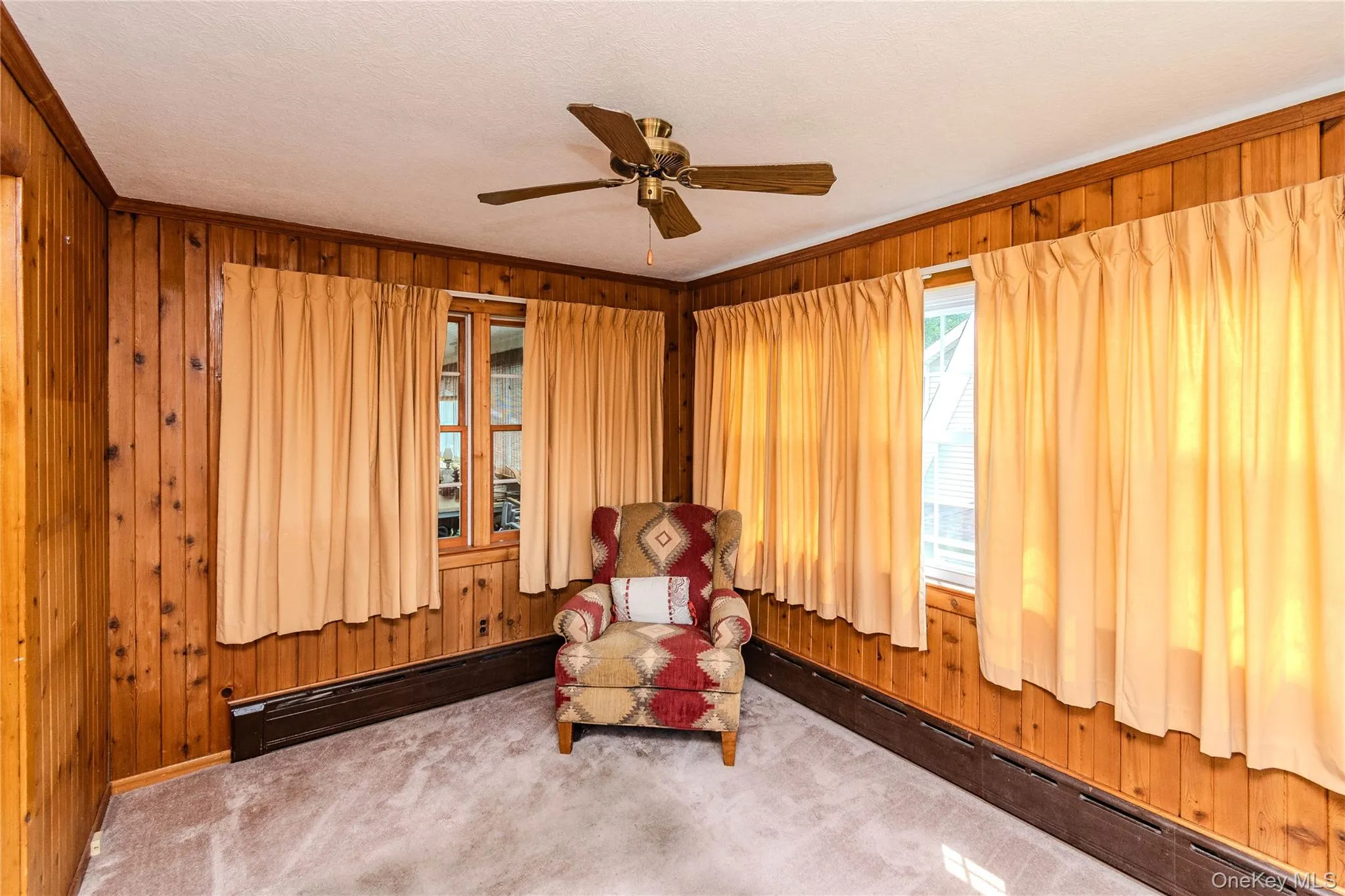 Room featuring wood paneling, multiple windows, a ceiling fan, and light-toned carpeting Room featuring wood paneling, multiple windows, a ceiling fan, and light-toned carpeting