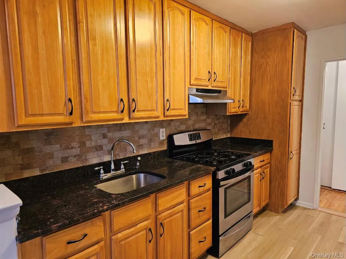 Kitchen featuring stainless steel gas range oven, dark stone countertops, light wood-type flooring, and brown cabinetry Kitchen featuring stainless steel gas range oven, dark stone countertops, light wood-type flooring, and brown cabinetry
