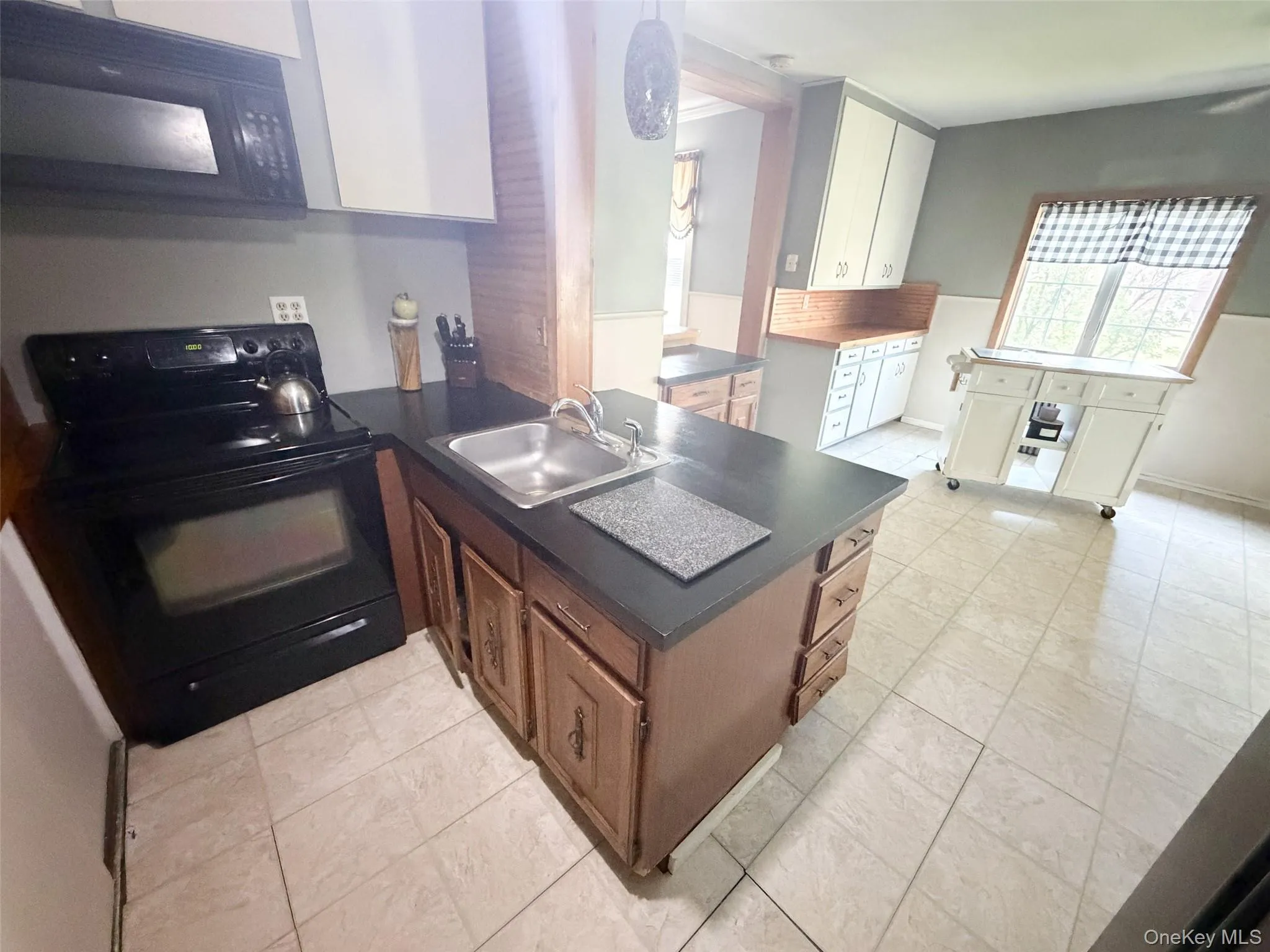 Kitchen featuring black appliances, wood-finish cabinetry, a stainless steel sink, dark countertops, and light-toned tile flooring Kitchen featuring black appliances, wood-finish cabinetry, a stainless steel sink, dark countertops, and light-toned tile flooring