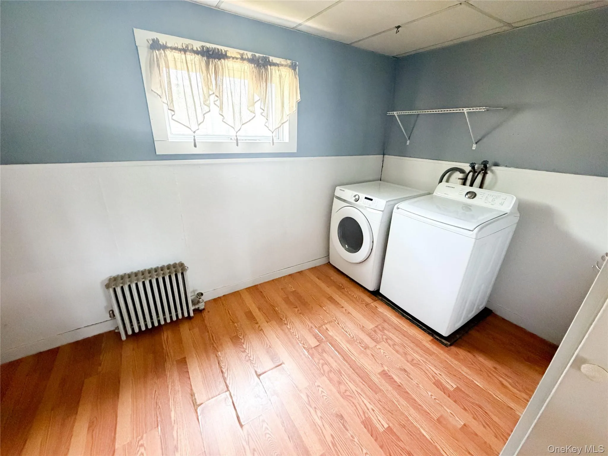 Dedicated laundry area featuring wood-finish flooring, a white baseboard, light blue walls, and an included radiator Dedicated laundry area featuring wood-finish flooring, a white baseboard, light blue walls, and an included radiator