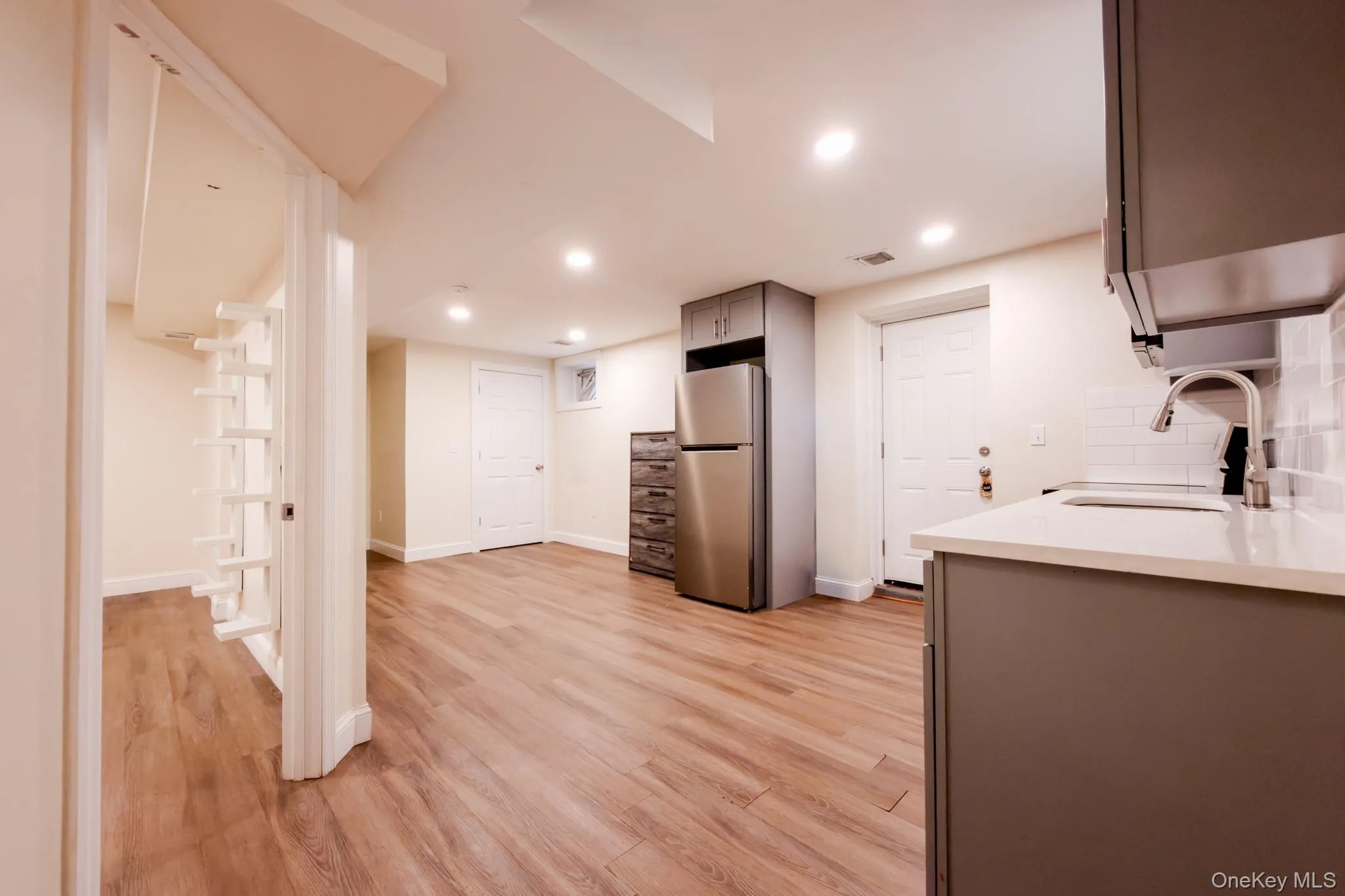 Open-concept room featuring wood-finish flooring, recessed lighting, and a kitchen area with gray cabinetry, white countertops, a stainless steel refrigerator, and a single-basin sink with a pull-down faucet Open-concept room featuring wood-finish flooring, recessed lighting, and a kitchen area with gray cabinetry, white countertops, a stainless steel refrigerator, and a single-basin sink with a pull-down faucet
