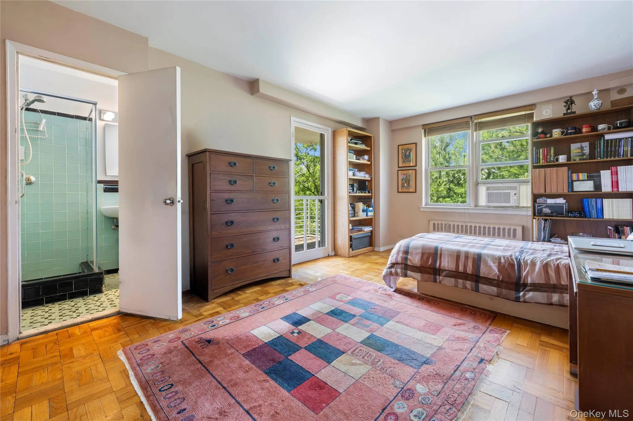 Hardwood parquet flooring throughout the room, featuring a built-in shelving unit, a window with exterior views, and a glass-paneled door leading to a balcony Hardwood parquet flooring throughout the room, featuring a built-in shelving unit, a window with exterior views, and a glass-paneled door leading to a balcony