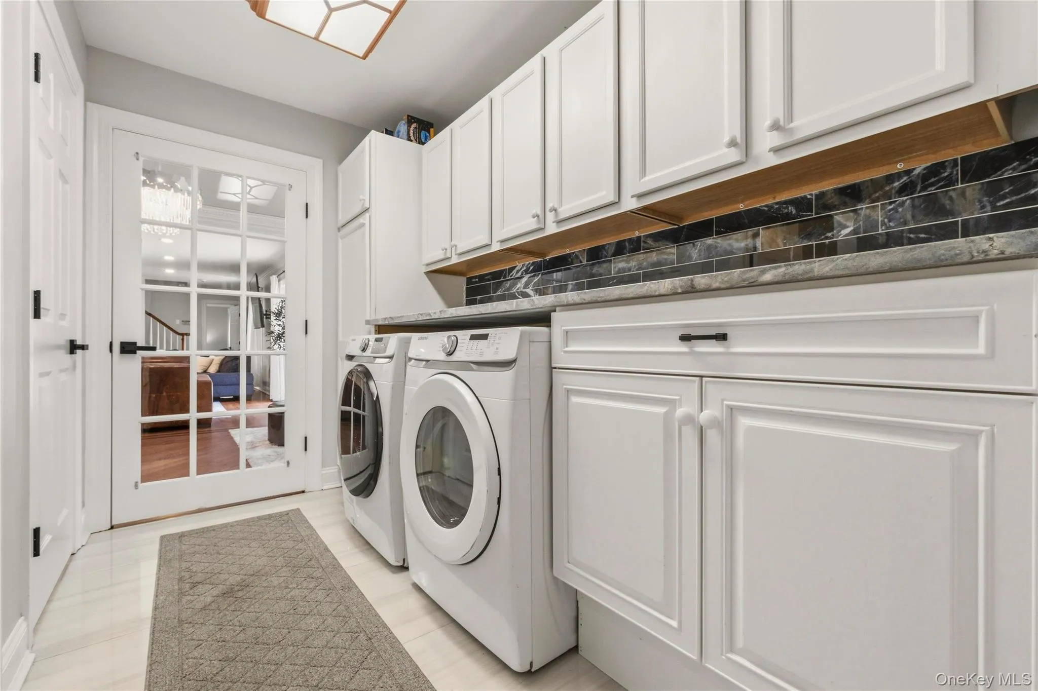 This is the first of 2 laundry rooms in the house. This beautifully designed laundry room offers both style and efficiency, featuring front-loading machines beneath a marble-patterned countertop and ample white cabinetry for storage. A striking marble tile backsplash adds a touch of sophistication, while a glass-paneled door opens to the adjacent living area. Bright, clean, and functional, this space makes everyday chores feel effortless. This is the first of 2 laundry rooms in the house. This beautifully designed laundry room offers both style and efficiency, featuring front-loading machines beneath a marble-patterned countertop and ample white cabinetry for storage. A striking marble tile backsplash adds a touch of sophistication, while a glass-paneled door opens to the adjacent living area. Bright, clean, and functional, this space makes everyday chores feel effortless.