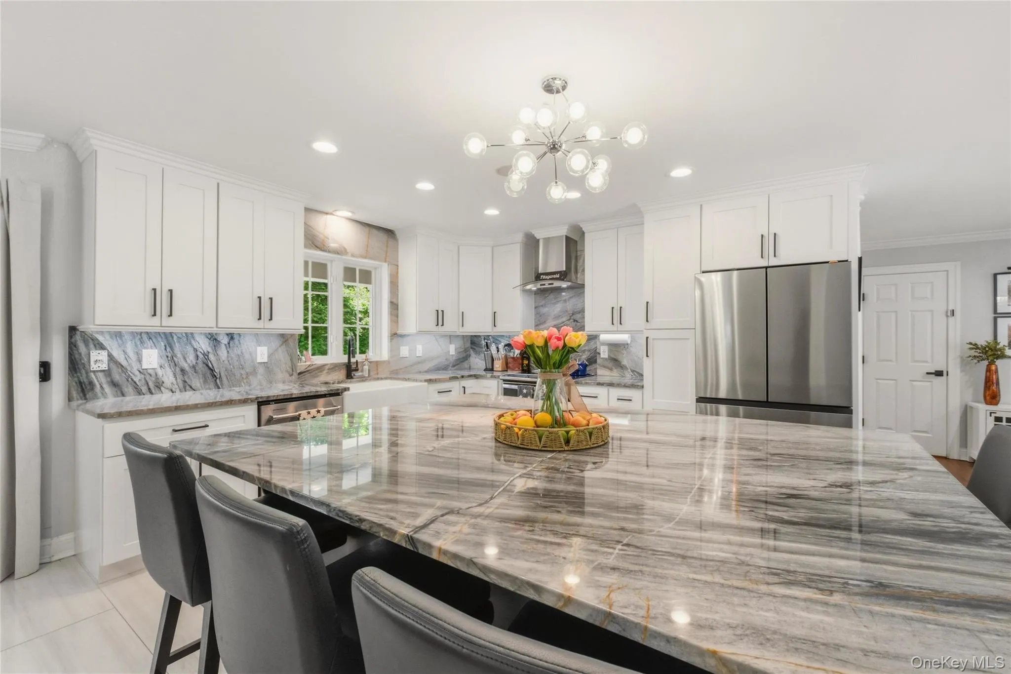 This stunning gourmet kitchen features a dramatic quartzite island with elegant veining, complemented by sleek white cabinetry and modern black hardware. Stainless steel appliances, a matching quartzite backsplash, and a contemporary chandelier elevate the space with a luxurious feel. Natural light pours in, perfect for cooking, gathering, and entertaining. This stunning gourmet kitchen features a dramatic quartzite island with elegant veining, complemented by sleek white cabinetry and modern black hardware. Stainless steel appliances, a matching quartzite backsplash, and a contemporary chandelier elevate the space with a luxurious feel. Natural light pours in, perfect for cooking, gathering, and entertaining.
