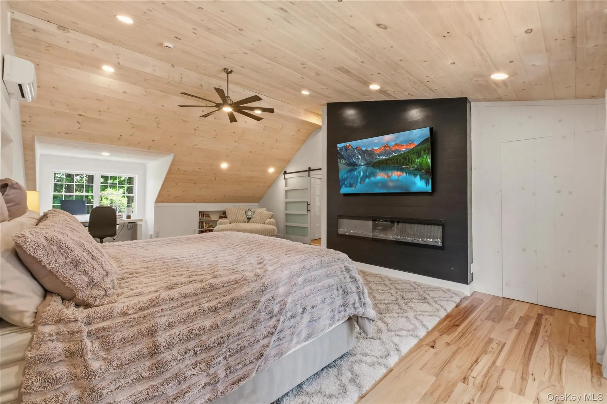 This inviting bedroom showcases a wood ceiling, built-in electric fireplace, and a sleek media wall for a luxurious feel. A reading nook and desk area add comfort and versatility to the space. This inviting bedroom showcases a wood ceiling, built-in electric fireplace, and a sleek media wall for a luxurious feel. A reading nook and desk area add comfort and versatility to the space.