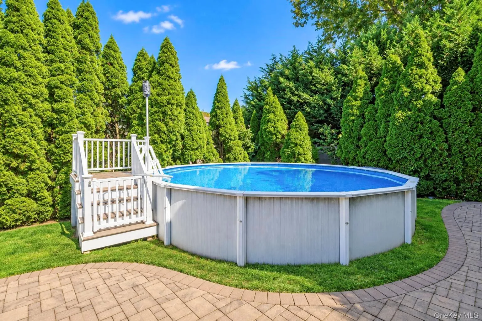 Above-ground pool with surrounding foliage, white wood-finish pool deck, and paver patio Above-ground pool with surrounding foliage, white wood-finish pool deck, and paver patio