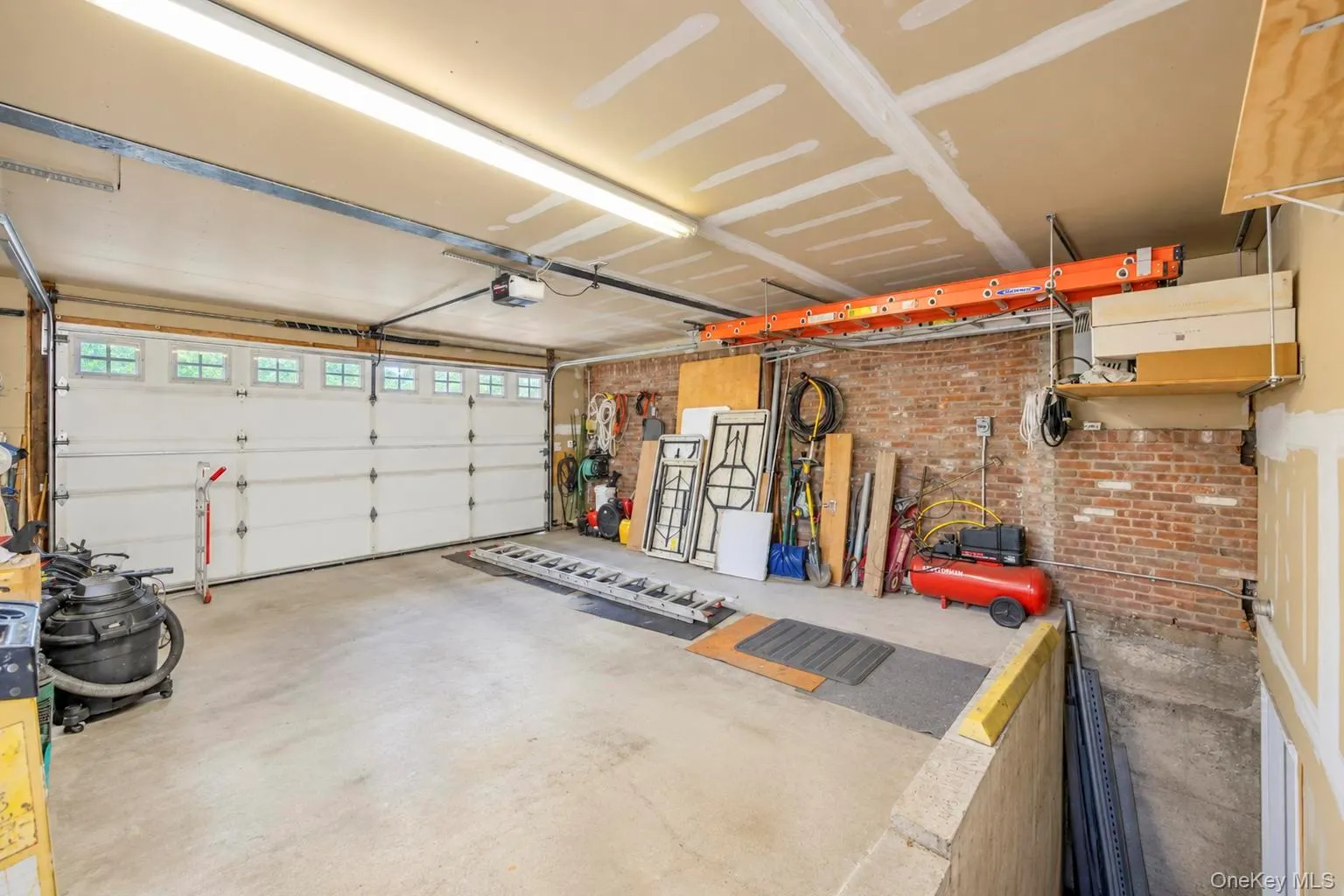 2 car garage, Concrete flooring, exposed brick wall, overhead lighting, segmented garage door with windows, and an overhead storage rack 2 car garage, Concrete flooring, exposed brick wall, overhead lighting, segmented garage door with windows, and an overhead storage rack
