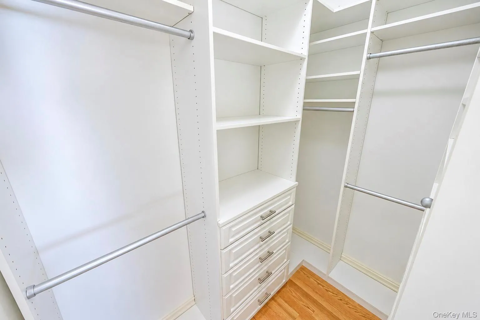 Custom closet system featuring multiple hanging rods, built-in shelving, integrated drawers with brushed nickel hardware, and wood-finish flooring Custom closet system featuring multiple hanging rods, built-in shelving, integrated drawers with brushed nickel hardware, and wood-finish flooring