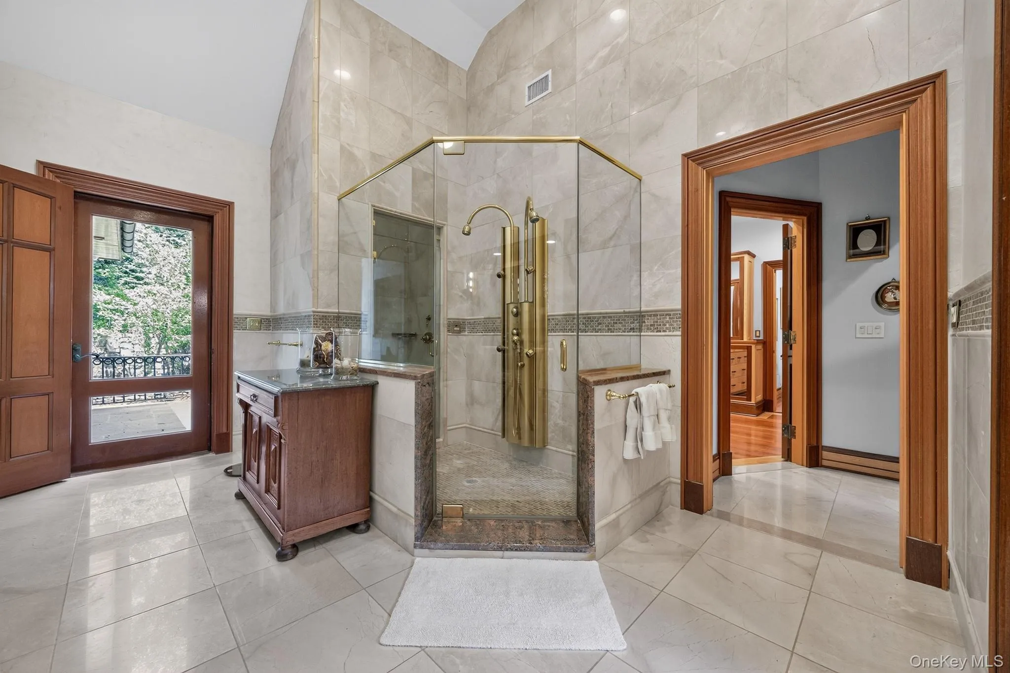 Glass-enclosed shower with multiple brass fixtures, stone tile walls and flooring, a wood vanity with a stone countertop, and a wood-framed door opening to an exterior balcony Glass-enclosed shower with multiple brass fixtures, stone tile walls and flooring, a wood vanity with a stone countertop, and a wood-framed door opening to an exterior balcony