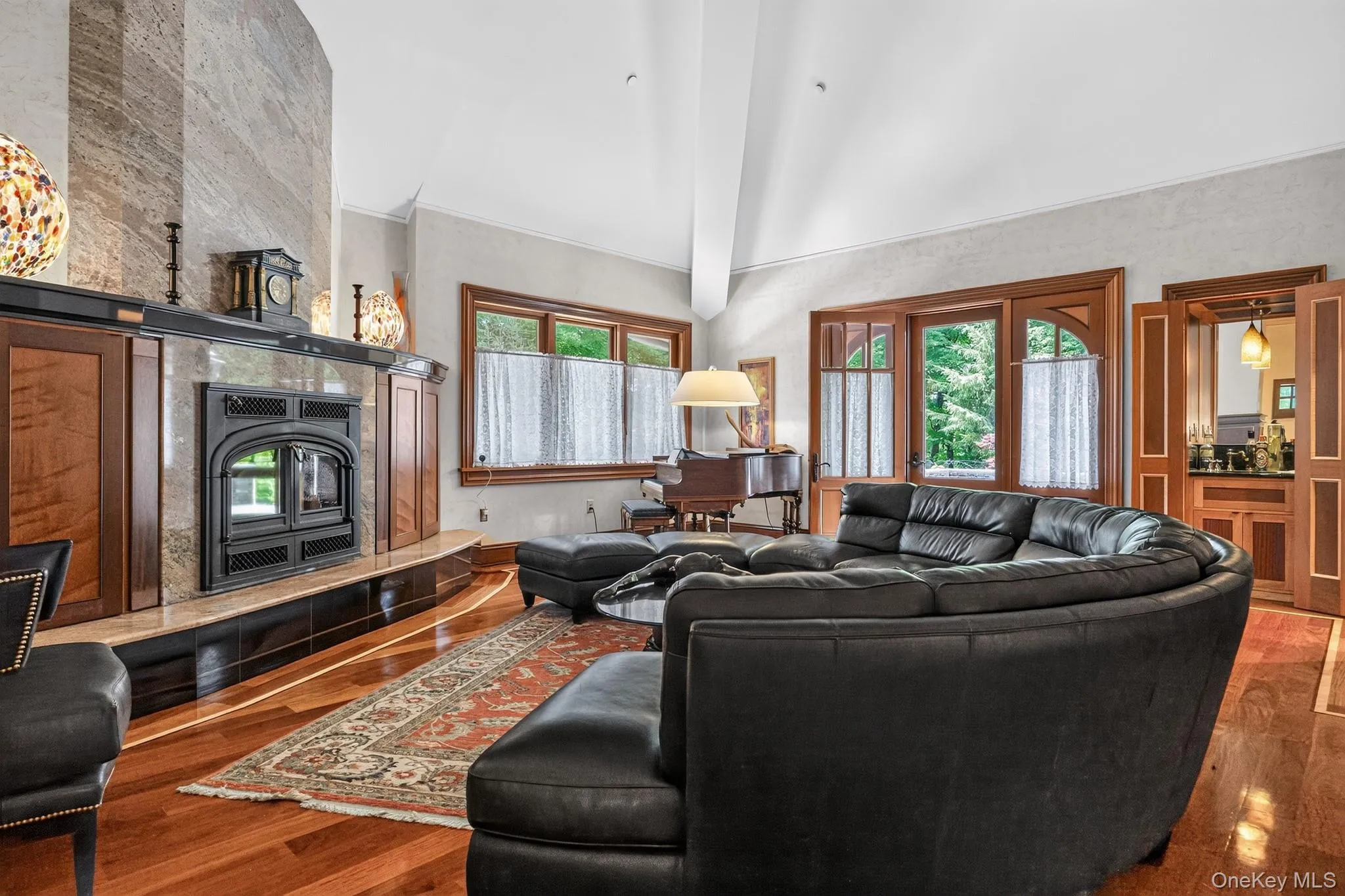 Spacious living area featuring a prominent fireplace with stone surround and wood mantel, rich wood-finish flooring, vaulted white ceiling, and arched wooden doors Spacious living area featuring a prominent fireplace with stone surround and wood mantel, rich wood-finish flooring, vaulted white ceiling, and arched wooden doors