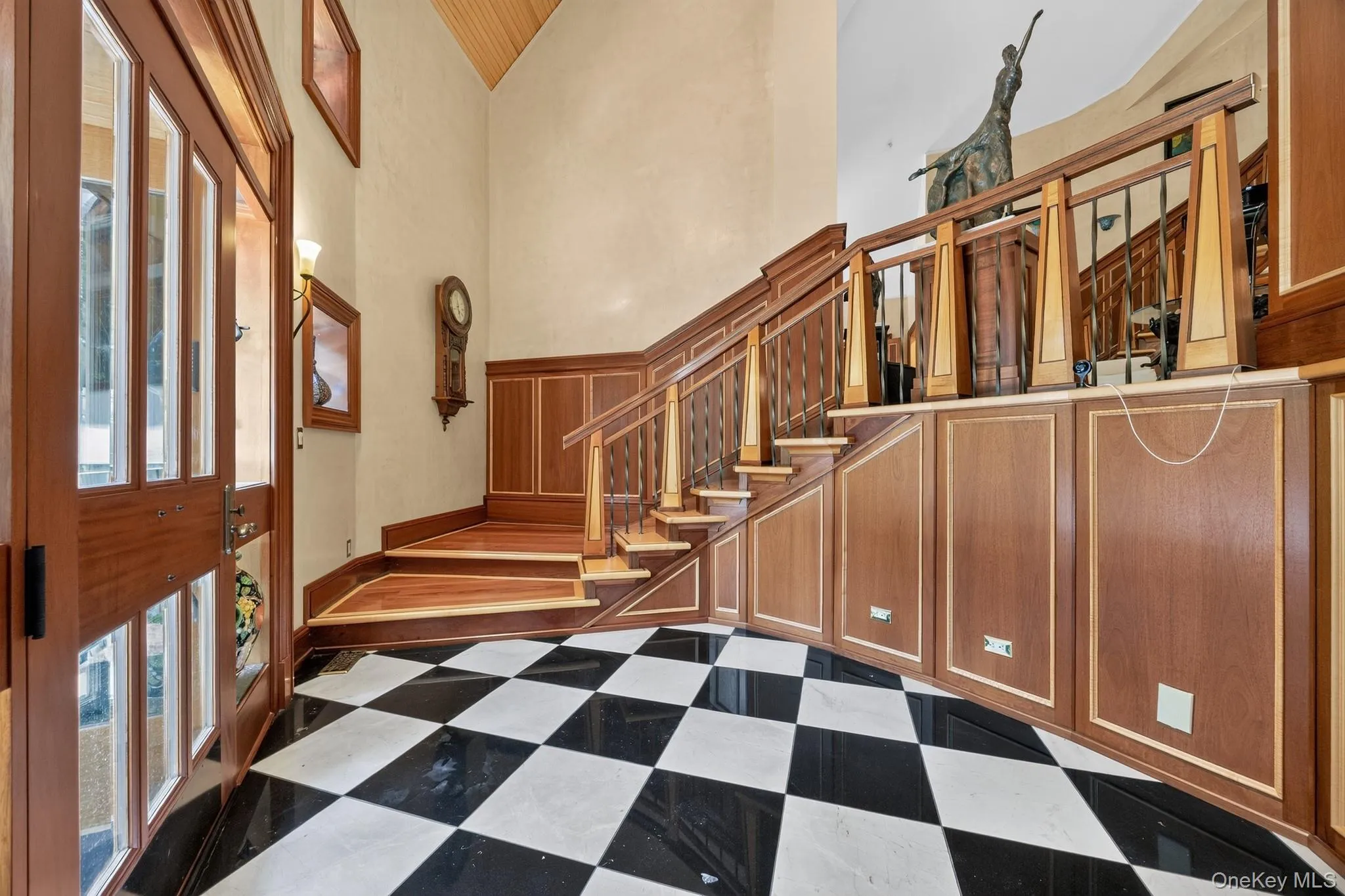 Foyer featuring black and white checkerboard flooring, a multi-paneled wood entry door, a sweeping wood staircase with decorative balusters, extensive wood paneling, and a vaulted wood-paneled ceiling Foyer featuring black and white checkerboard flooring, a multi-paneled wood entry door, a sweeping wood staircase with decorative balusters, extensive wood paneling, and a vaulted wood-paneled ceiling