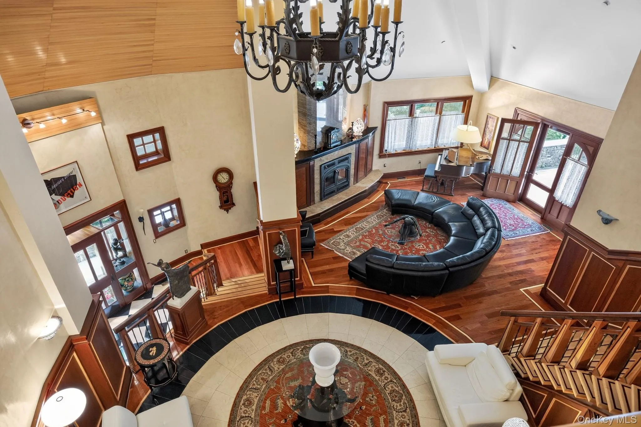 Two-story living area featuring a prominent dark-toned chandelier, a fireplace with a stone surround, and wood-finish flooring with an inlaid circular tile design Two-story living area featuring a prominent dark-toned chandelier, a fireplace with a stone surround, and wood-finish flooring with an inlaid circular tile design