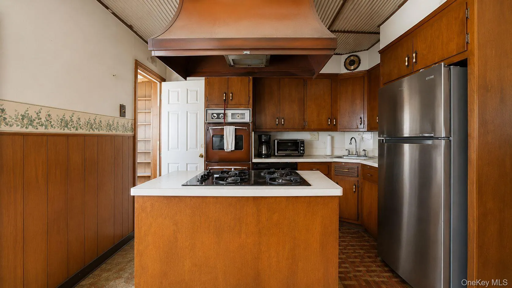 Kitchen featuring wood cabinetry, stainless steel refrigerator, white countertops, built-in oven, and a center island with cooktop Kitchen featuring wood cabinetry, stainless steel refrigerator, white countertops, built-in oven, and a center island with cooktop