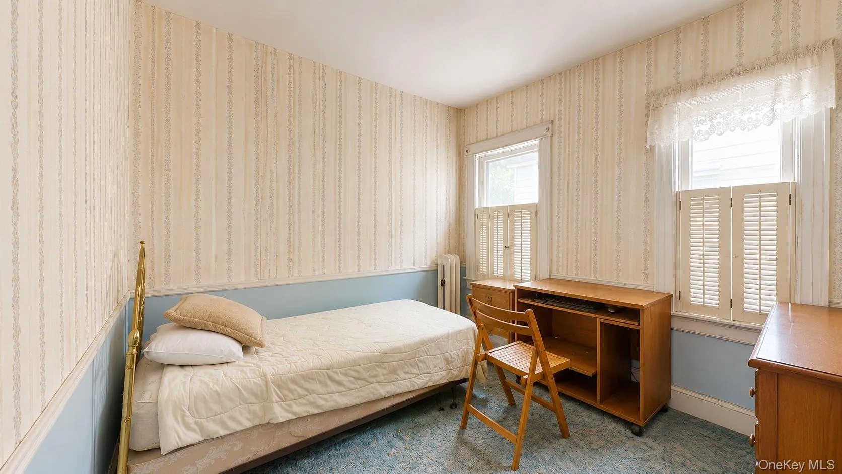 Bedroom featuring two windows with white louvered shutters, a built-in radiator, and light-toned patterned wallpaper with a blue wainscoting detail Bedroom featuring two windows with white louvered shutters, a built-in radiator, and light-toned patterned wallpaper with a blue wainscoting detail