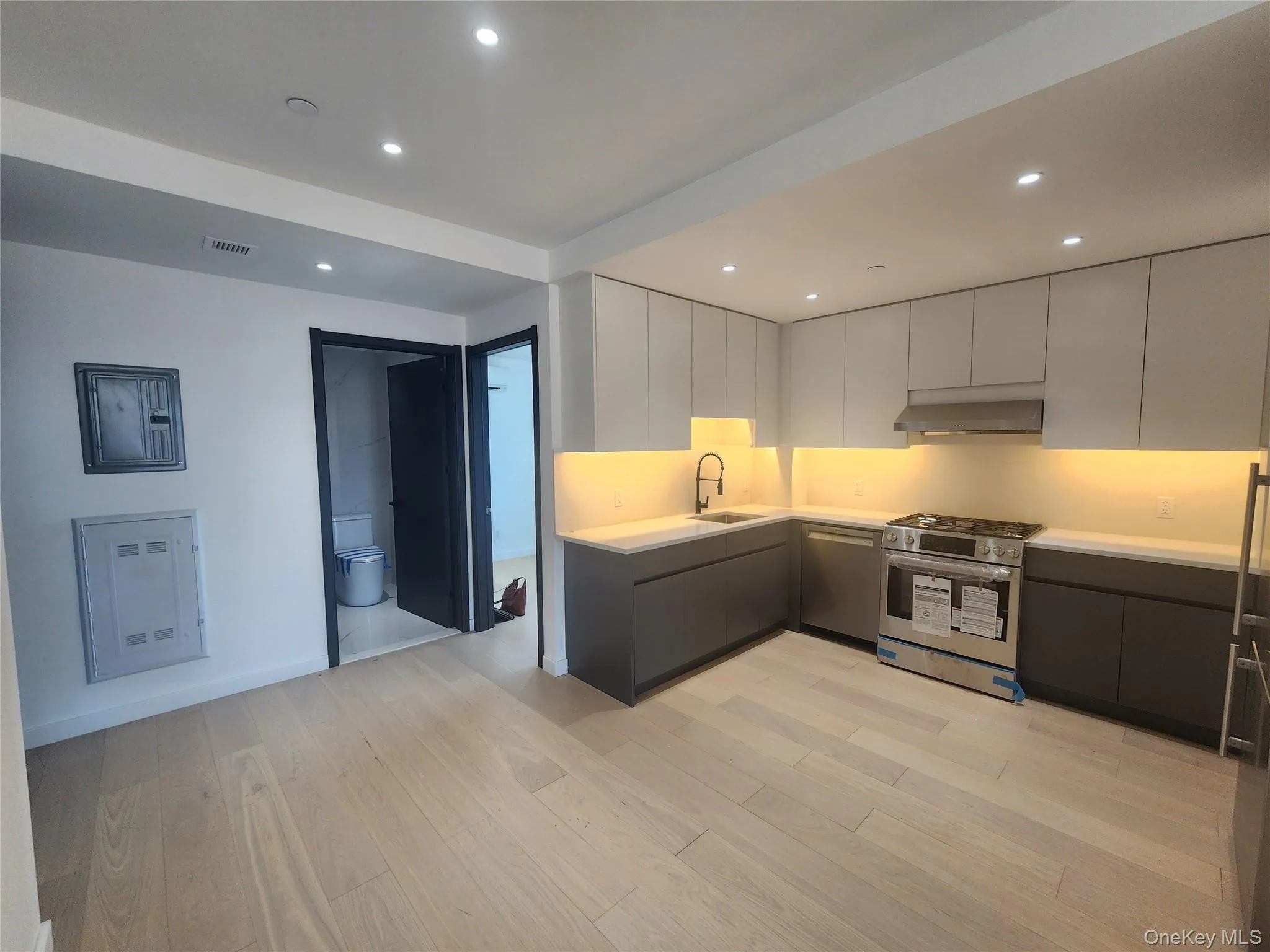 Modern kitchen featuring light wood-finish flooring, white flat-panel upper cabinetry, dark flat-panel lower cabinetry, stainless steel appliances, and integrated under-cabinet lighting Modern kitchen featuring light wood-finish flooring, white flat-panel upper cabinetry, dark flat-panel lower cabinetry, stainless steel appliances, and integrated under-cabinet lighting