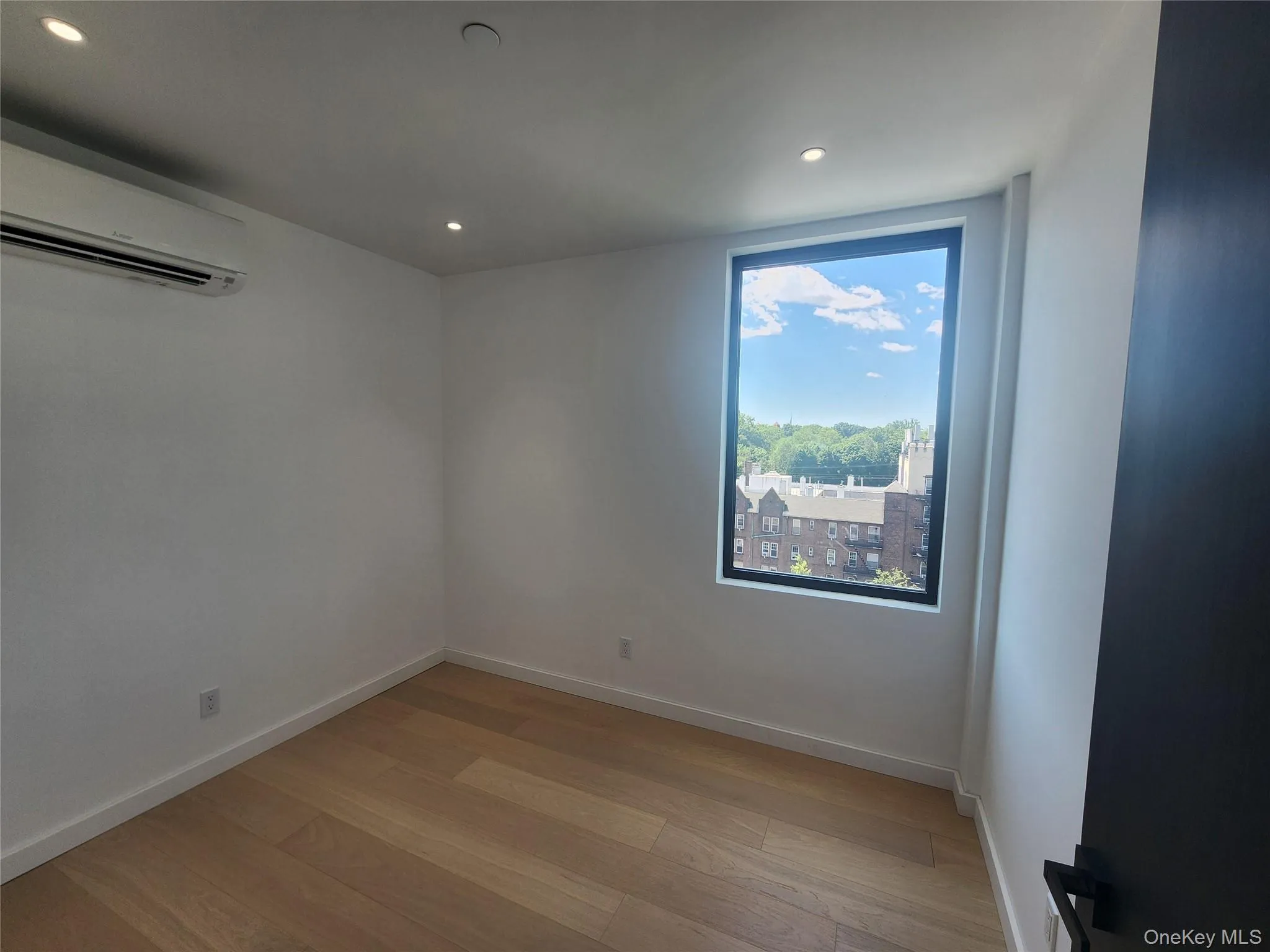 Bright room featuring light wood-finish flooring, recessed ceiling lighting, a large window with a black frame, and a wall-mounted air conditioning unit Bright room featuring light wood-finish flooring, recessed ceiling lighting, a large window with a black frame, and a wall-mounted air conditioning unit