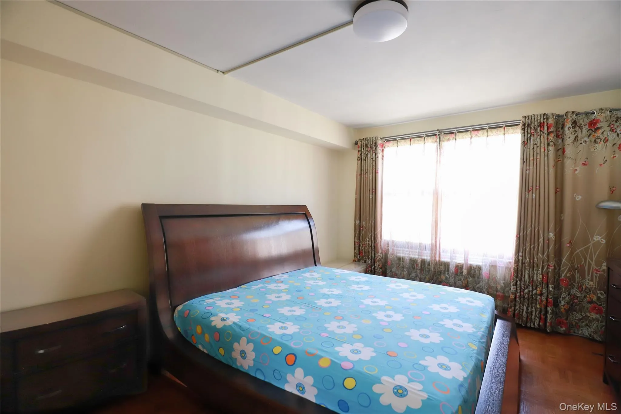 Bright interior bedroom featuring a large window with sheers and patterned drapes, wood-finish flooring, and overhead lighting Bright interior bedroom featuring a large window with sheers and patterned drapes, wood-finish flooring, and overhead lighting