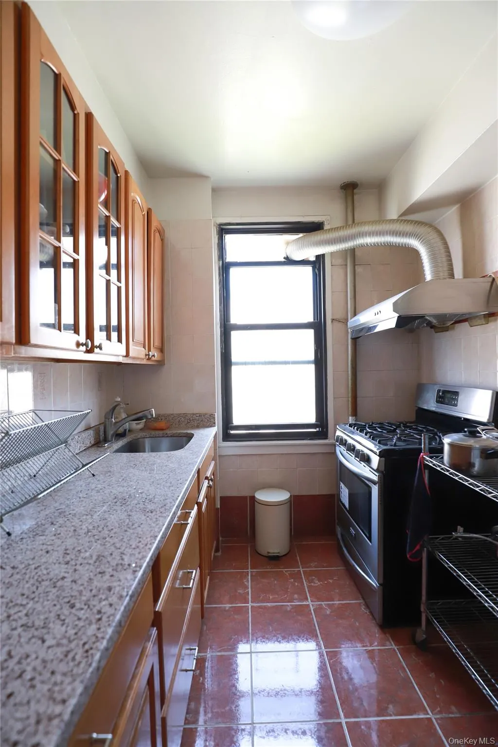 Galley kitchen featuring wood-finish cabinetry, granite-style countertops, stainless steel sink, tile flooring, and a stainless steel gas range with exhaust hood Galley kitchen featuring wood-finish cabinetry, granite-style countertops, stainless steel sink, tile flooring, and a stainless steel gas range with exhaust hood
