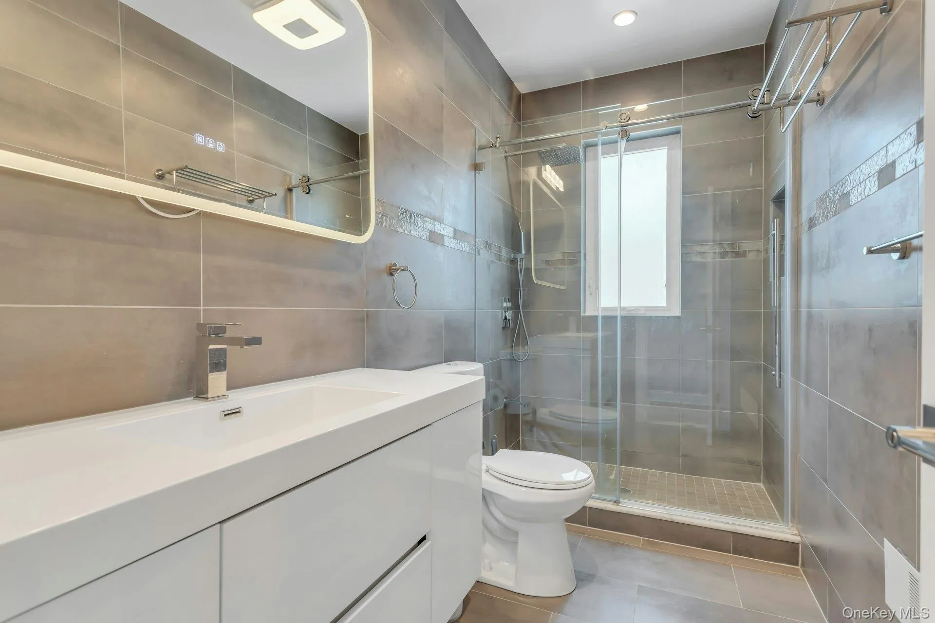 Modern bathroom featuring a white vanity with an integrated sink, a frameless lighted, anti fog mirror, and a glass-enclosed shower with porcelain tile design Modern bathroom featuring a white vanity with an integrated sink, a frameless lighted, anti fog mirror, and a glass-enclosed shower with porcelain tile design