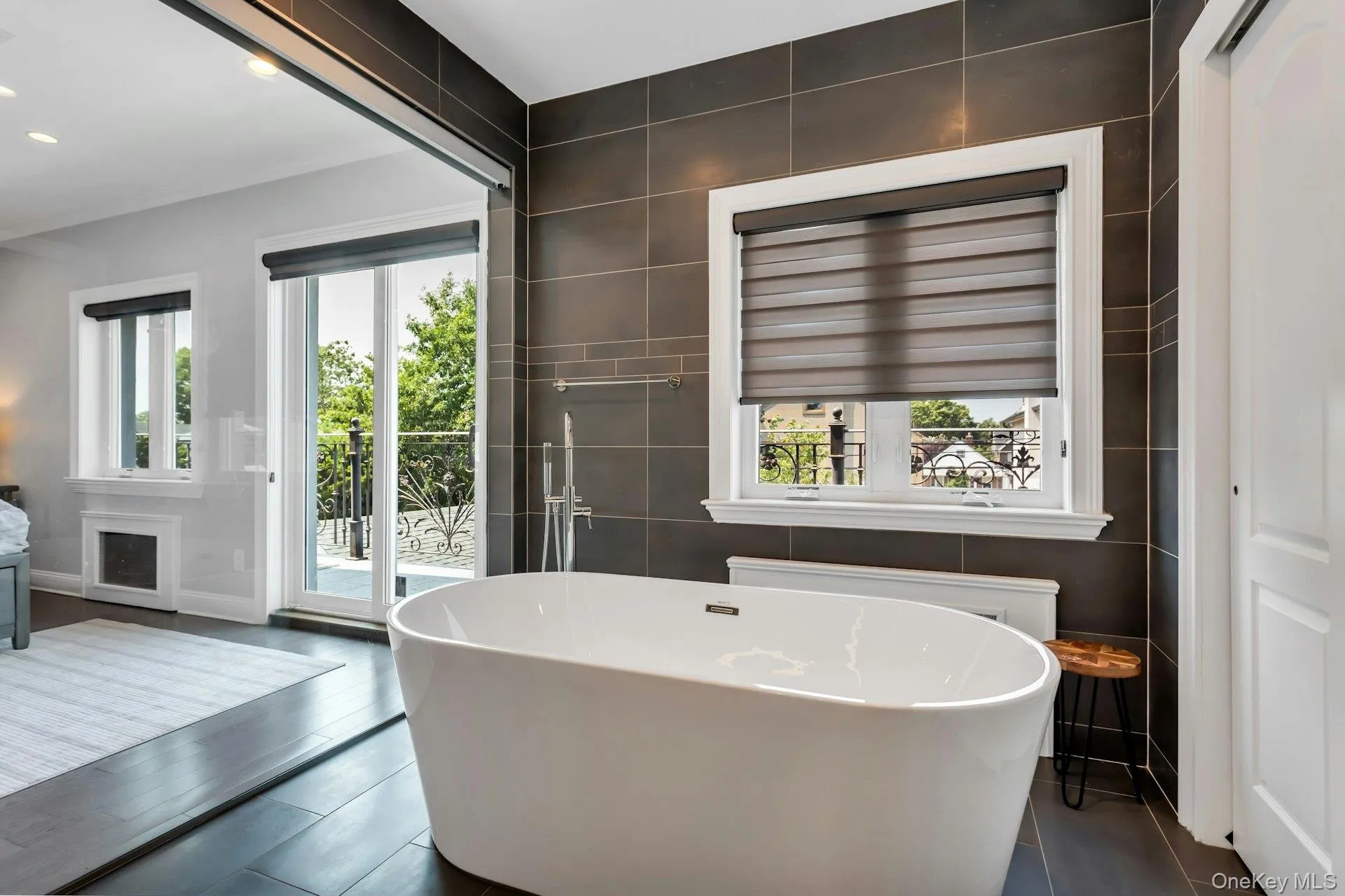 Architectural Digest! Freestanding soaking tub with floor-mounted faucet, dark large-format tile accent wall, dark wood-finish flooring, and a glass-paneled balcony access Architectural Digest! Freestanding soaking tub with floor-mounted faucet, dark large-format tile accent wall, dark wood-finish flooring, and a glass-paneled balcony access