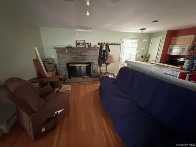 Wood-finish flooring, stone-clad fireplace with black insert, light green wall paint, kitchen with white built-in microwave and range, and light-toned upper cabinetry Wood-finish flooring, stone-clad fireplace with black insert, light green wall paint, kitchen with white built-in microwave and range, and light-toned upper cabinetry