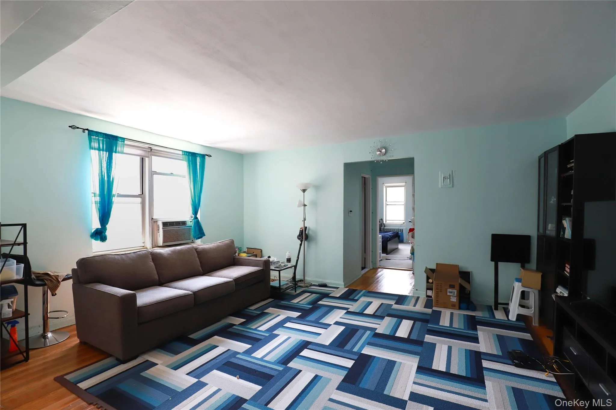 Bright living area featuring wood-finish flooring, light blue wall paint, and a large window with an integrated air conditioning unit Bright living area featuring wood-finish flooring, light blue wall paint, and a large window with an integrated air conditioning unit