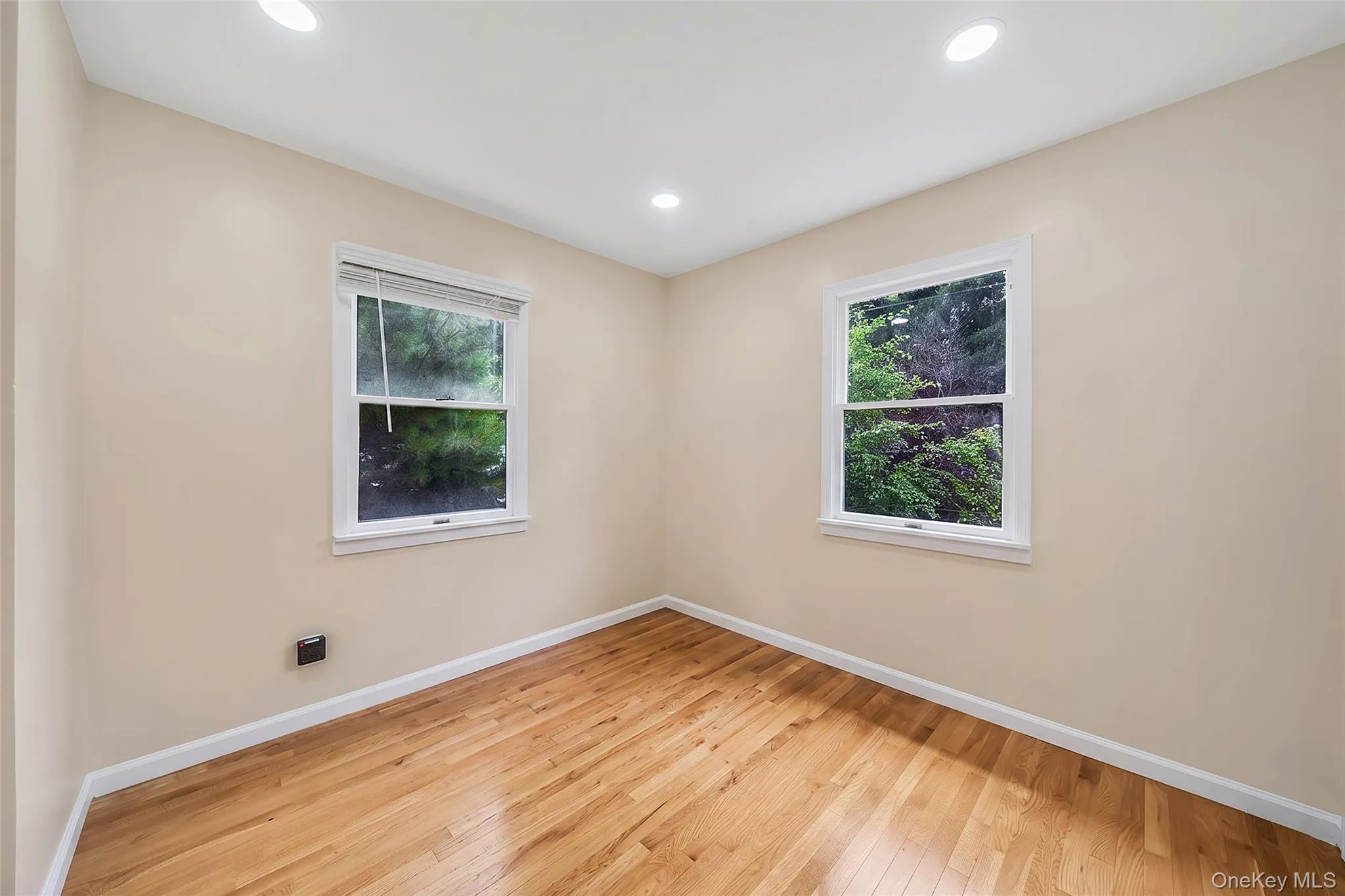 Room featuring two double-hung windows with white trim, light beige wall paint, white baseboards, wood-finish flooring, and recessed ceiling lighting Room featuring two double-hung windows with white trim, light beige wall paint, white baseboards, wood-finish flooring, and recessed ceiling lighting