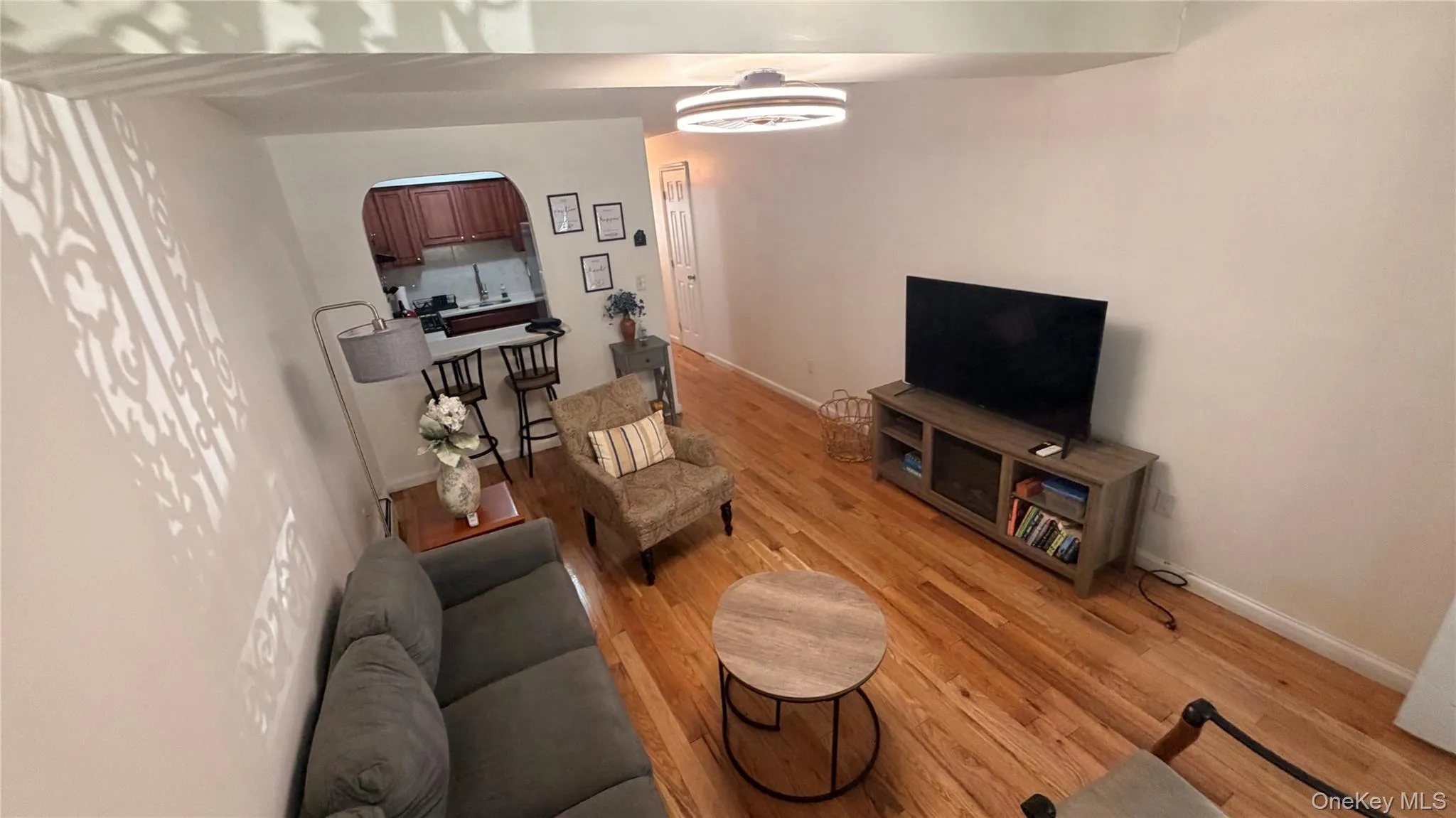 Hardwood flooring throughout a living area featuring a contemporary ceiling light fixture and an arched doorway leading to a kitchen with dark wood-finish cabinetry Hardwood flooring throughout a living area featuring a contemporary ceiling light fixture and an arched doorway leading to a kitchen with dark wood-finish cabinetry