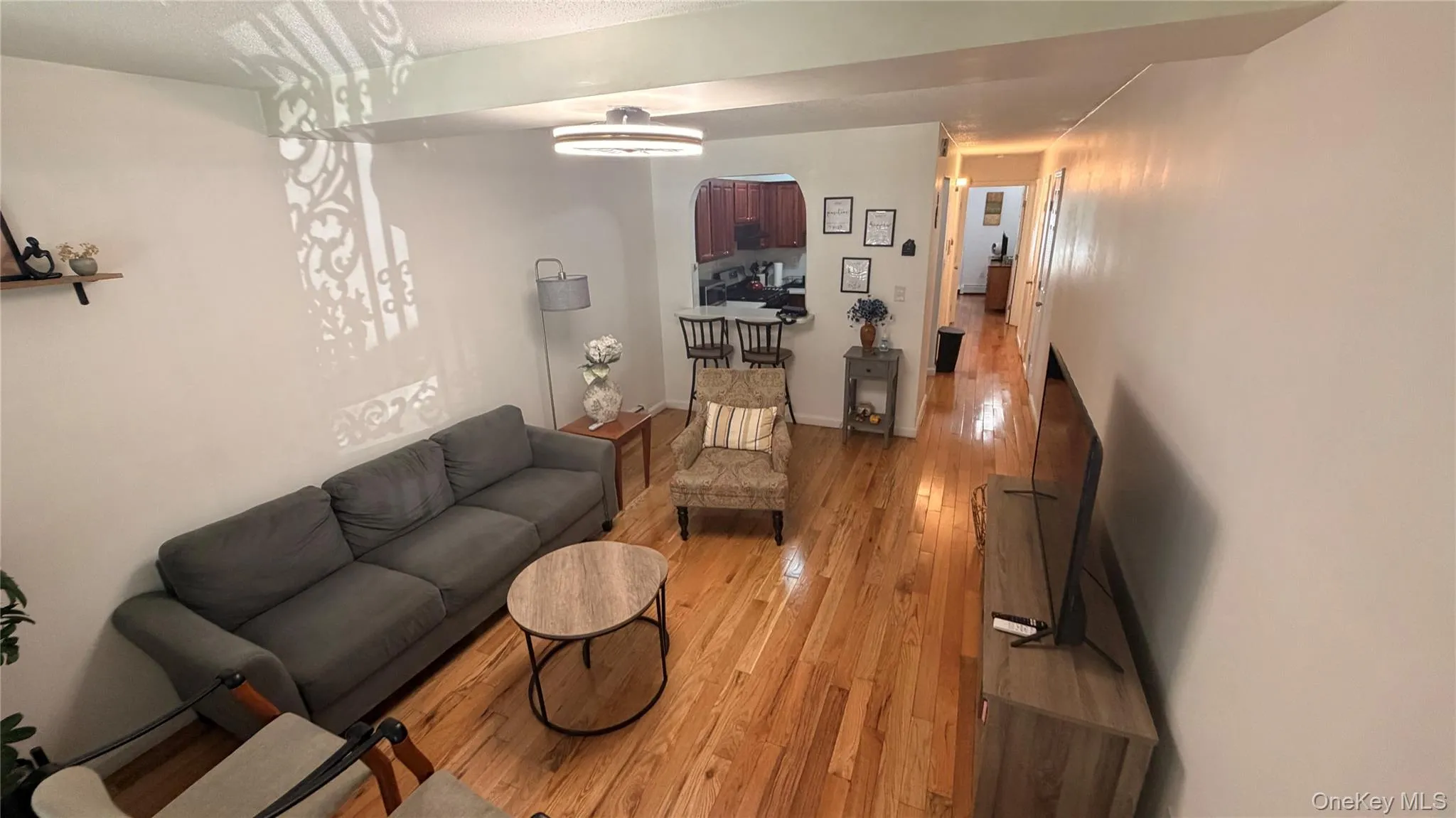 Hardwood flooring extends throughout this expansive living area, featuring a contemporary ceiling light fixture, a pass-through opening to the kitchen, and a hallway with additional lighting Hardwood flooring extends throughout this expansive living area, featuring a contemporary ceiling light fixture, a pass-through opening to the kitchen, and a hallway with additional lighting