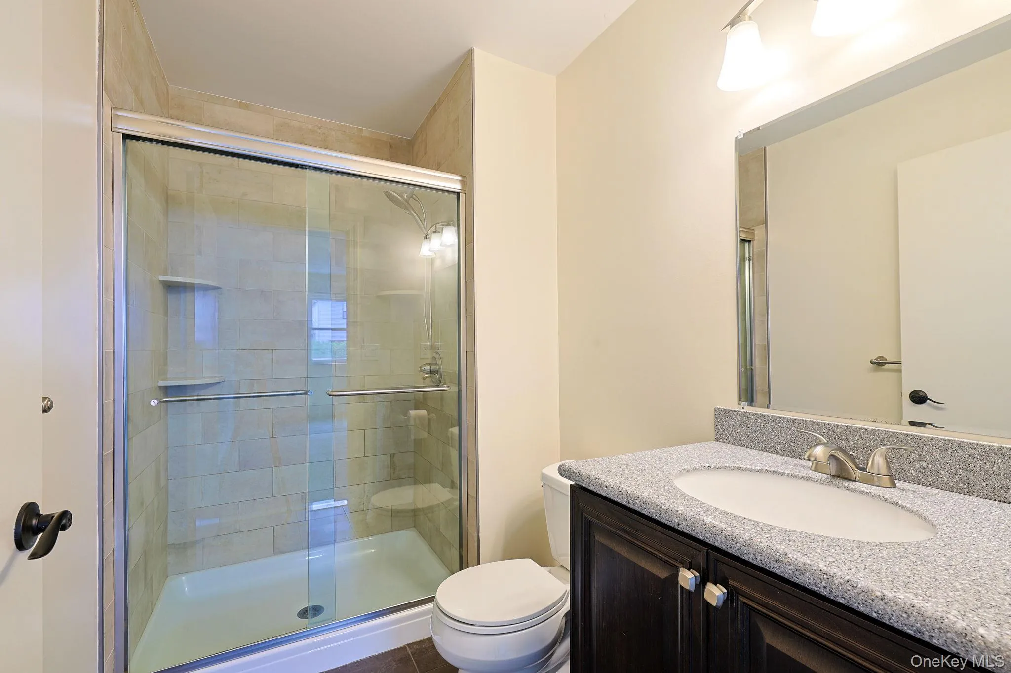 Bathroom featuring a glass-enclosed shower with tiled walls and built-in shelving, a dark wood-finish vanity with a speckled countertop and integrated sink, a wall-mounted mirror, and a three-light fixture Bathroom featuring a glass-enclosed shower with tiled walls and built-in shelving, a dark wood-finish vanity with a speckled countertop and integrated sink, a wall-mounted mirror, and a three-light fixture
