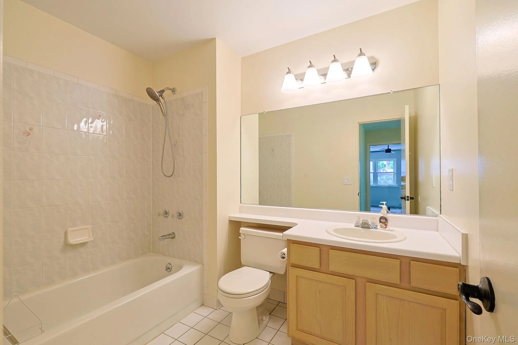 Full hallway bathroom featuring a white tub with tile surround, a single vanity with wood-finish cabinetry, a large mirror, and overhead vanity lighting Full hallway bathroom featuring a white tub with tile surround, a single vanity with wood-finish cabinetry, a large mirror, and overhead vanity lighting