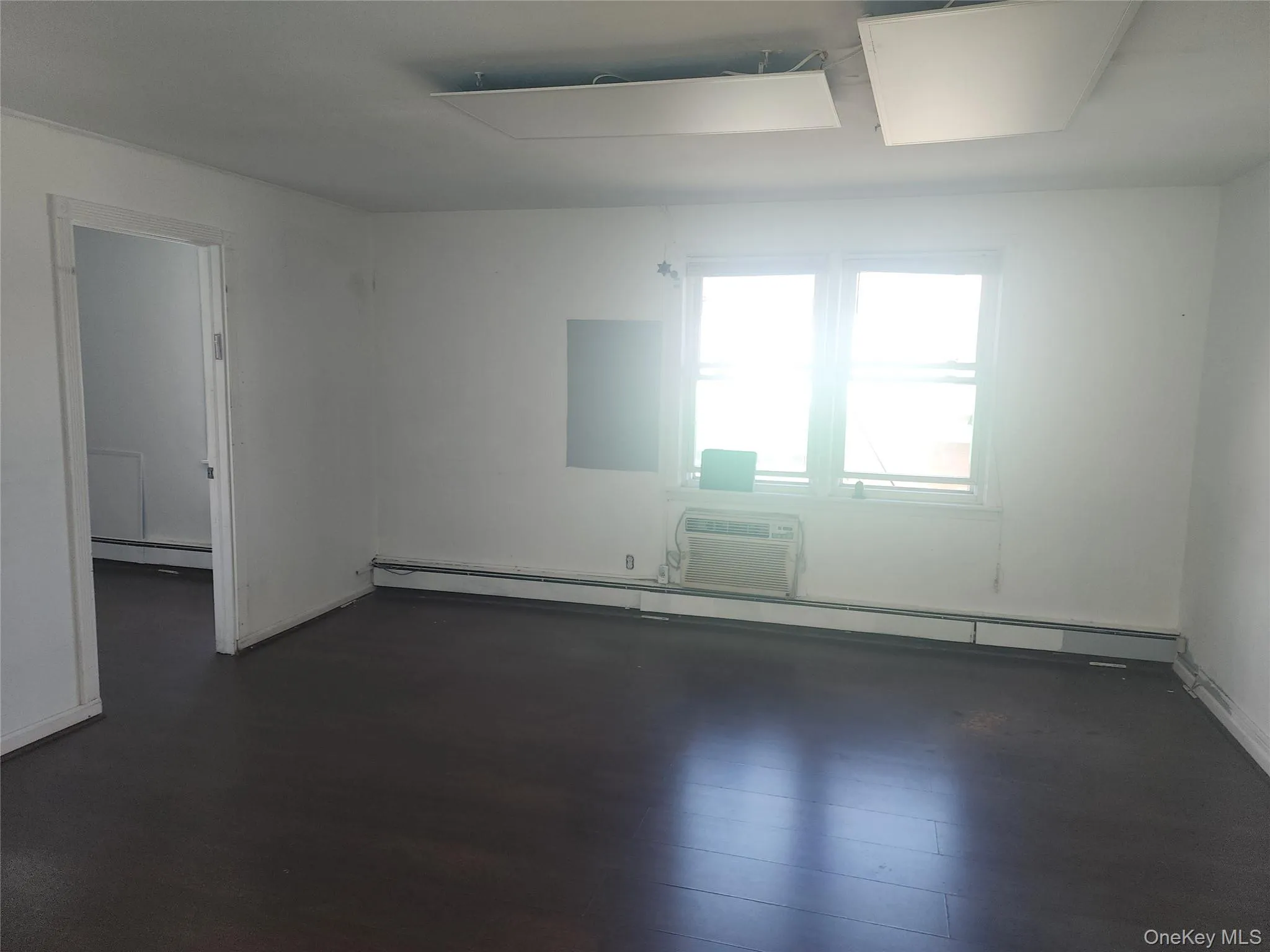 Room with dark wood-finish flooring, white walls, and a window with a wall-mounted air conditioning unit Room with dark wood-finish flooring, white walls, and a window with a wall-mounted air conditioning unit