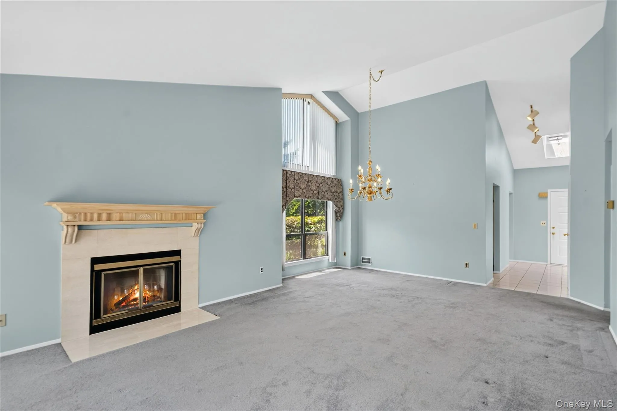 Spacious living area featuring a central fireplace with a light-toned mantel and hearth, vaulted ceilings, and traditional chandelier Spacious living area featuring a central fireplace with a light-toned mantel and hearth, vaulted ceilings, and traditional chandelier
